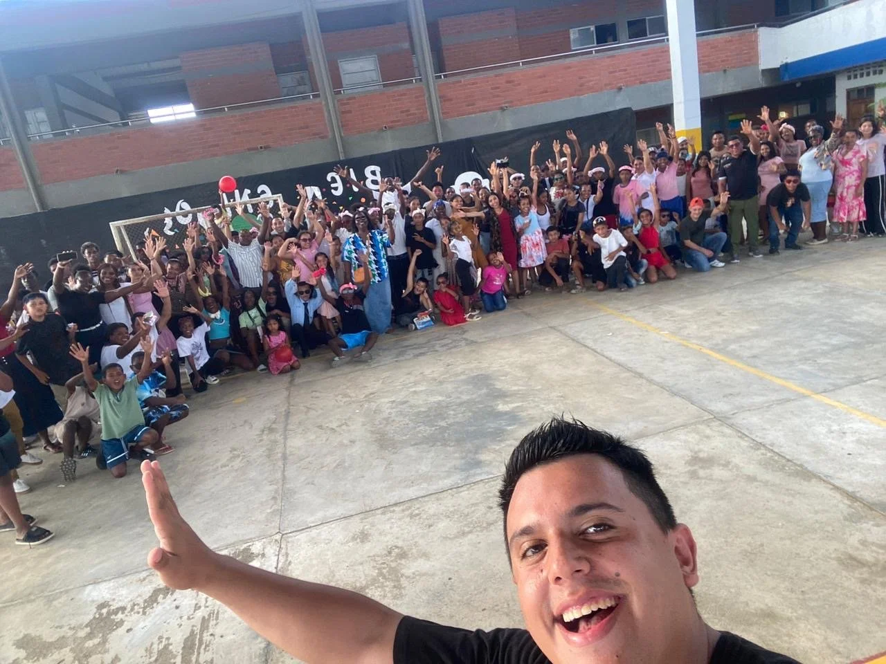 A man taking a selfie with a large group of children and adults gathered behind him, all smiling and waving. The setting is an indoor or covered outdoor space with a brick building in the background.
