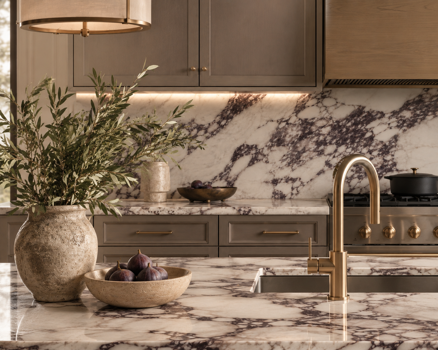 Kitchen with marble countertop, backsplash, grey cabinets, and gold faucet. Contains a plant in a beige vase, a bowl of figs, and a pot on the stove.