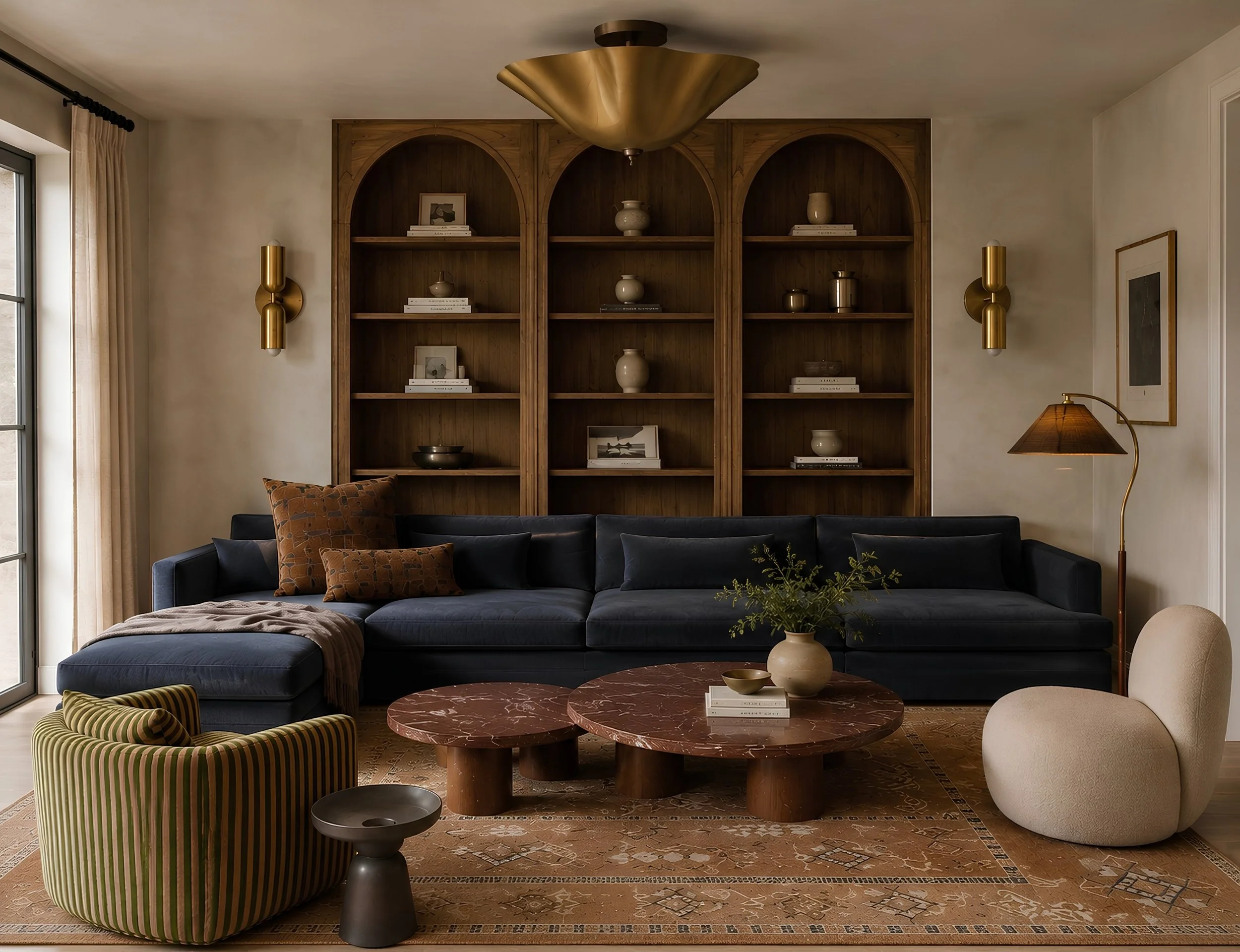 Living room with a dark blue sectional sofa, round marble coffee tables, a velvet green armchair with striped pattern, and a beige blanket on the sofa. Behind are wooden built-in shelves with decorative vases, books, and framed artwork. Gold wall sconces and a floor lamp with a gold arc stand on either side of the shelves. There is a window with beige curtains on the left.