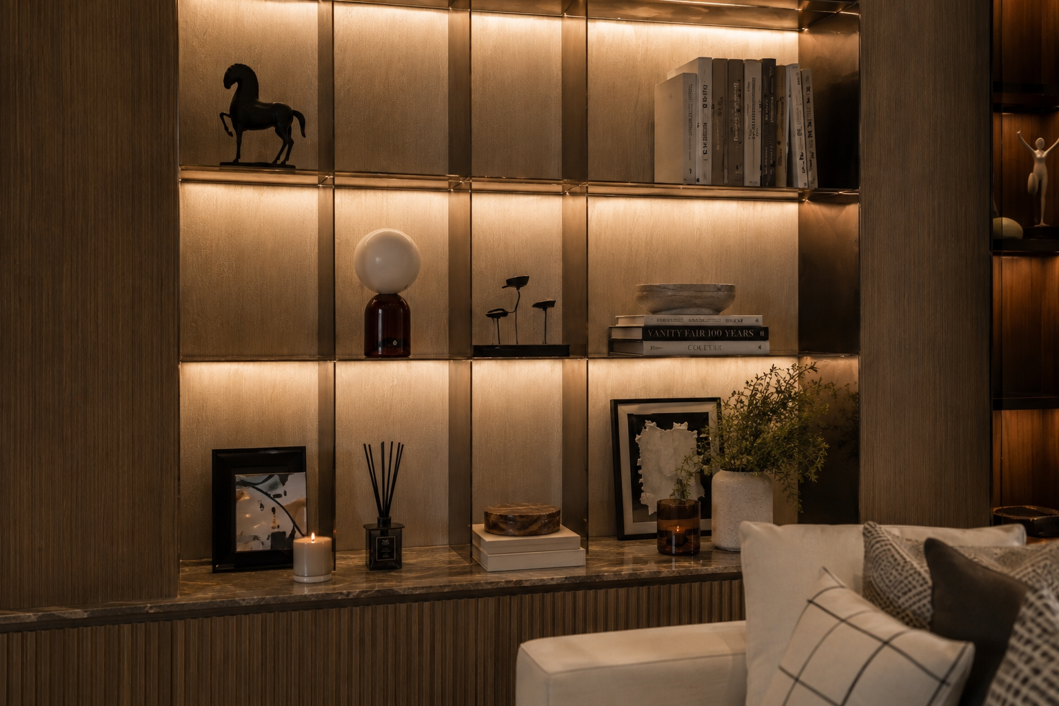 Wooden wall with illuminated open shelves displaying books, decorative objects, candles, picture frames, and plants in a modern living room.