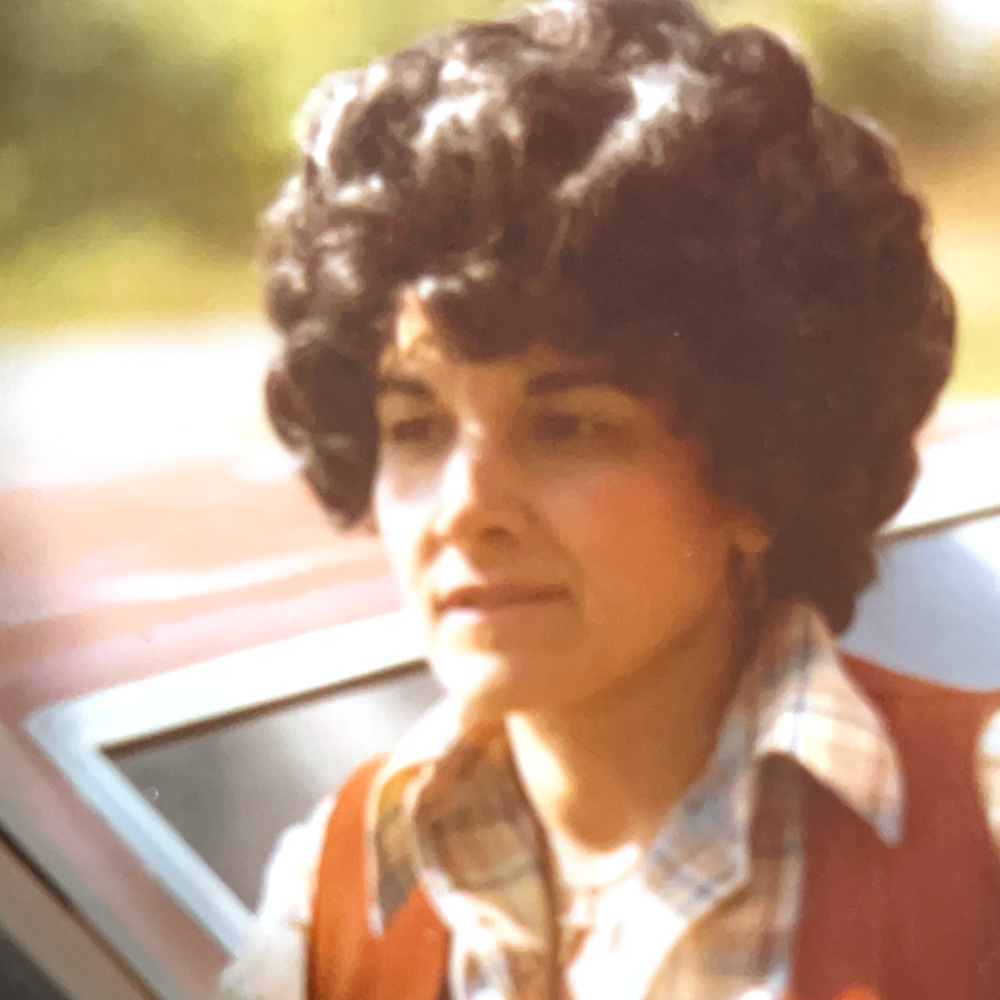 A woman, Carmela, with short curly hair wearing a plaid shirt and red sweater looks out the window with a thoughtful expression.