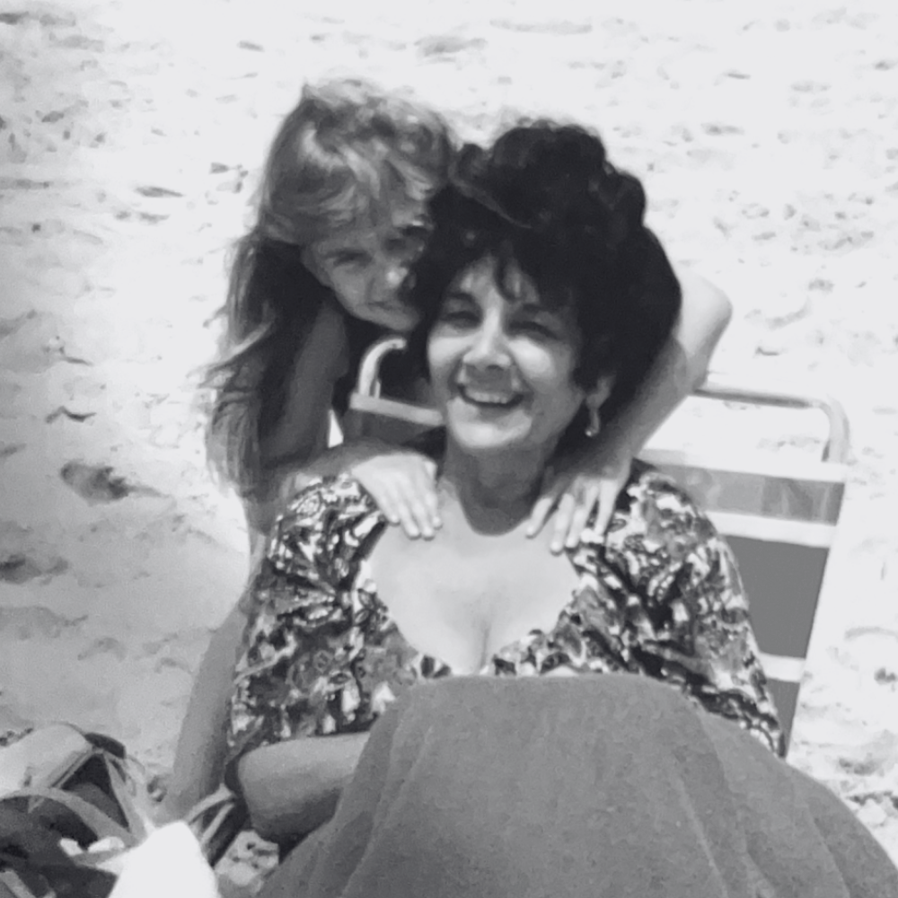Carmela and Nicole smiling and hugging at the beach, one seated in a towel or chair and the other leaning over her from behind.