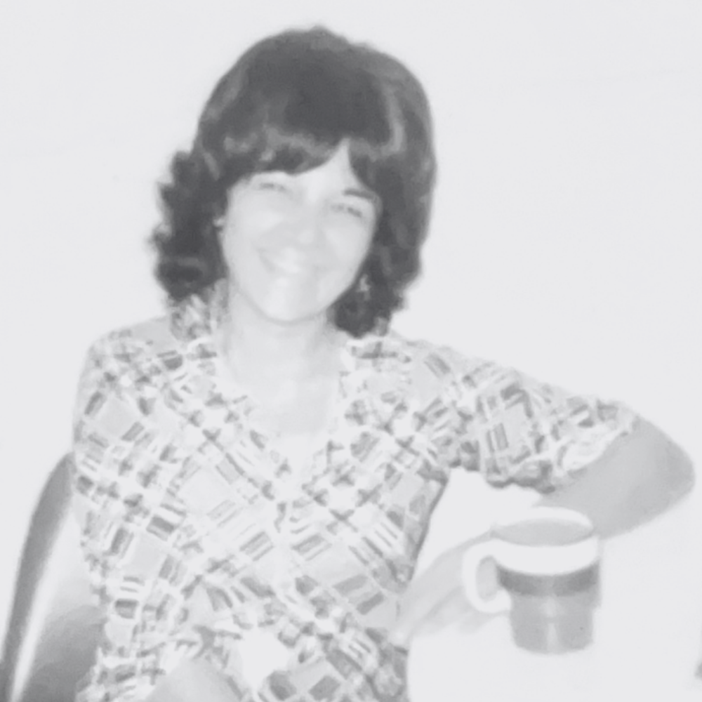 Black and white photo of Carmela smiling with a coffee mug in her hand, sitting at a table.