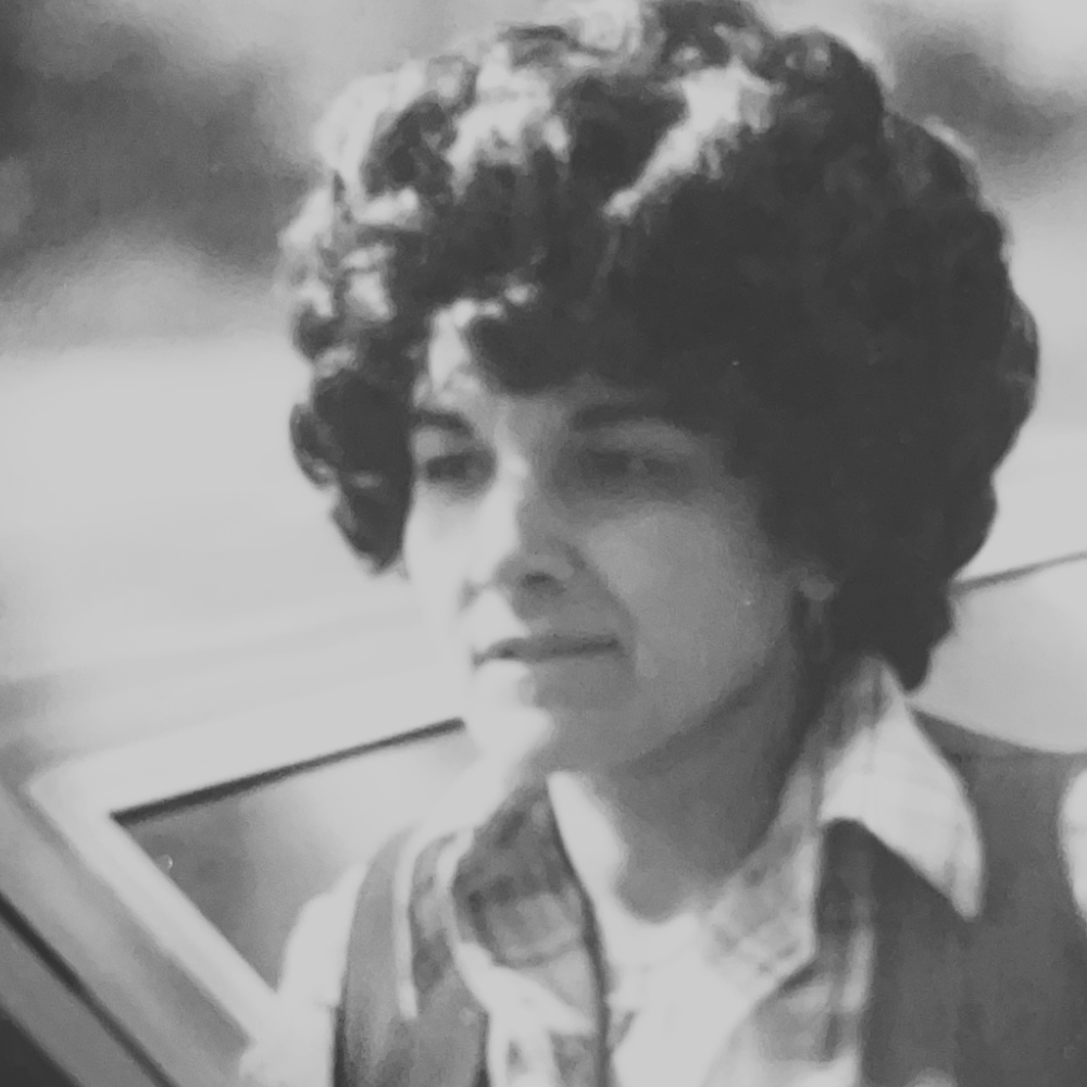 Black and white photograph of Carmela with short, curly hair, wearing a collared shirt and a jacket, sitting outdoors next to a car.