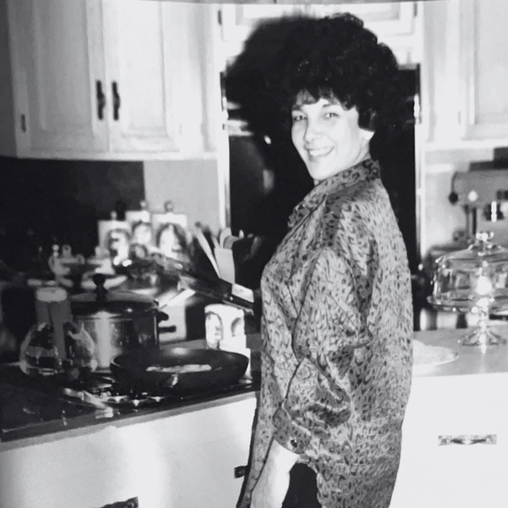 Carmela smiling in a kitchen, standing next to a stove with some pots and kitchen items on the counter.