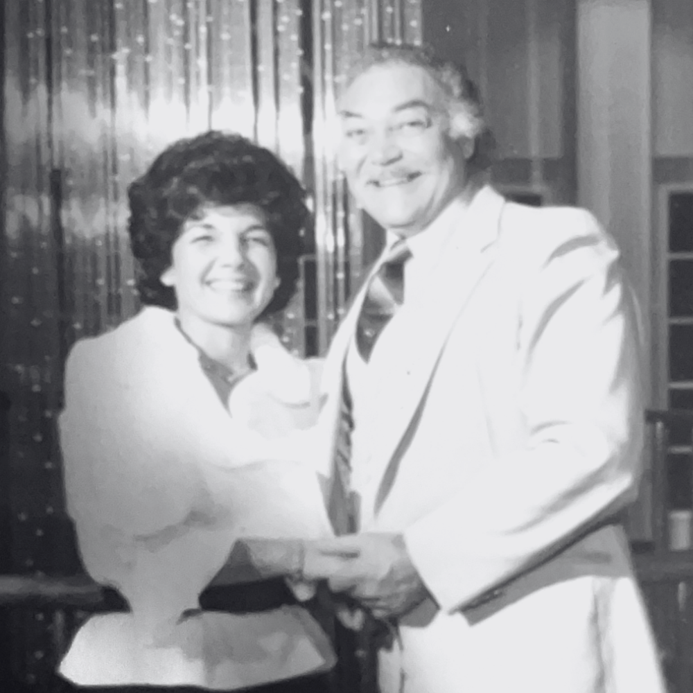 A black and white photo of Carmela and her husband holding hands, dressed in light-colored suits, standing indoors with a festive background.