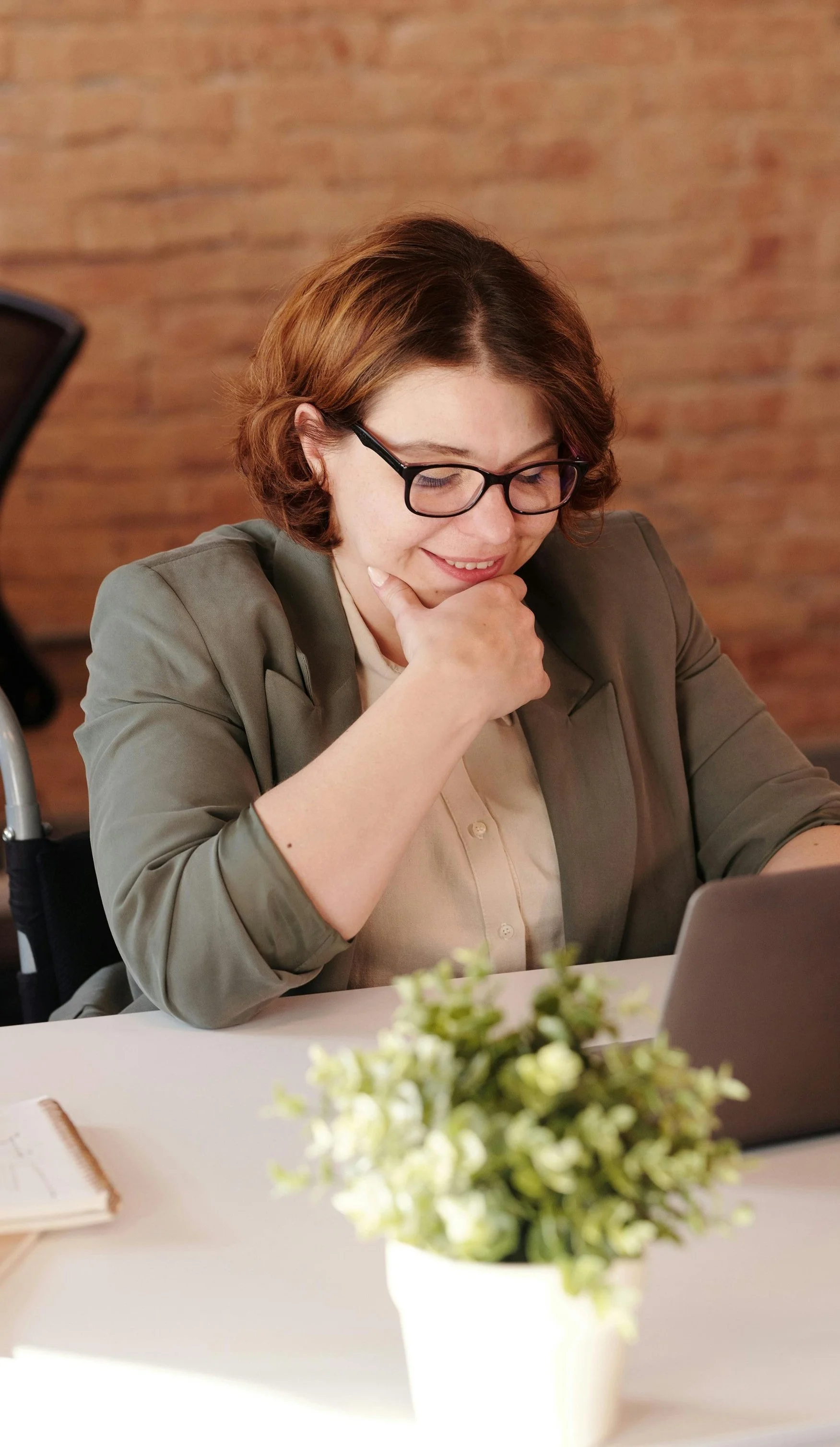 A woman with glasses and short brown hair, wearing a gray blazer and a beige shirt, sitting at a table, smiling while looking at her laptop with a potted plant and notebook on the table.