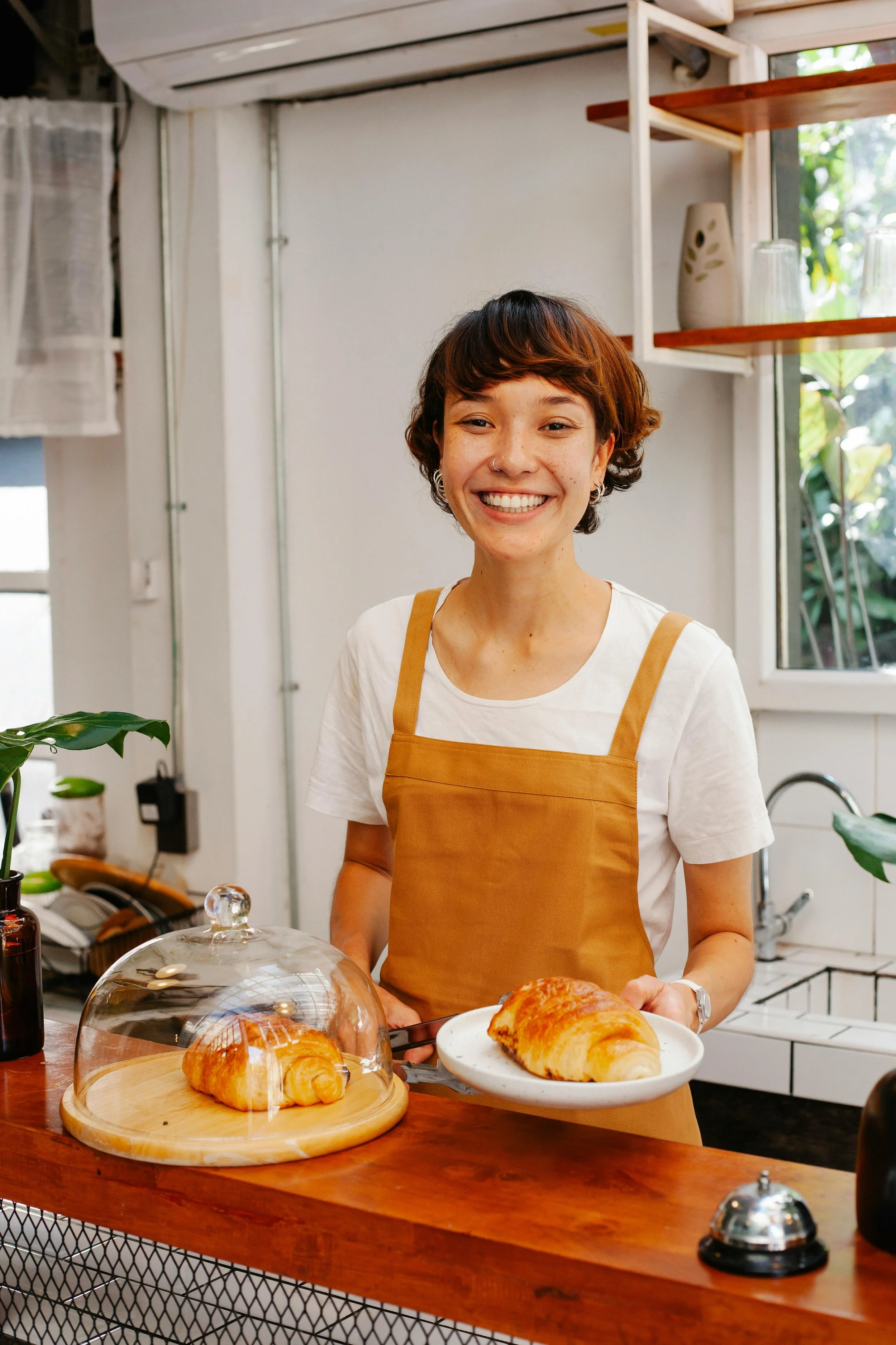 A smiling woman wearing a white t-shirt and an orange apron standing behind a wooden counter in a cozy bakery or cafe, holding a plate with a croissant. There is another croissant under a glass dome on the counter. The background has a white wall, wooden shelves, and a window with greenery outside.