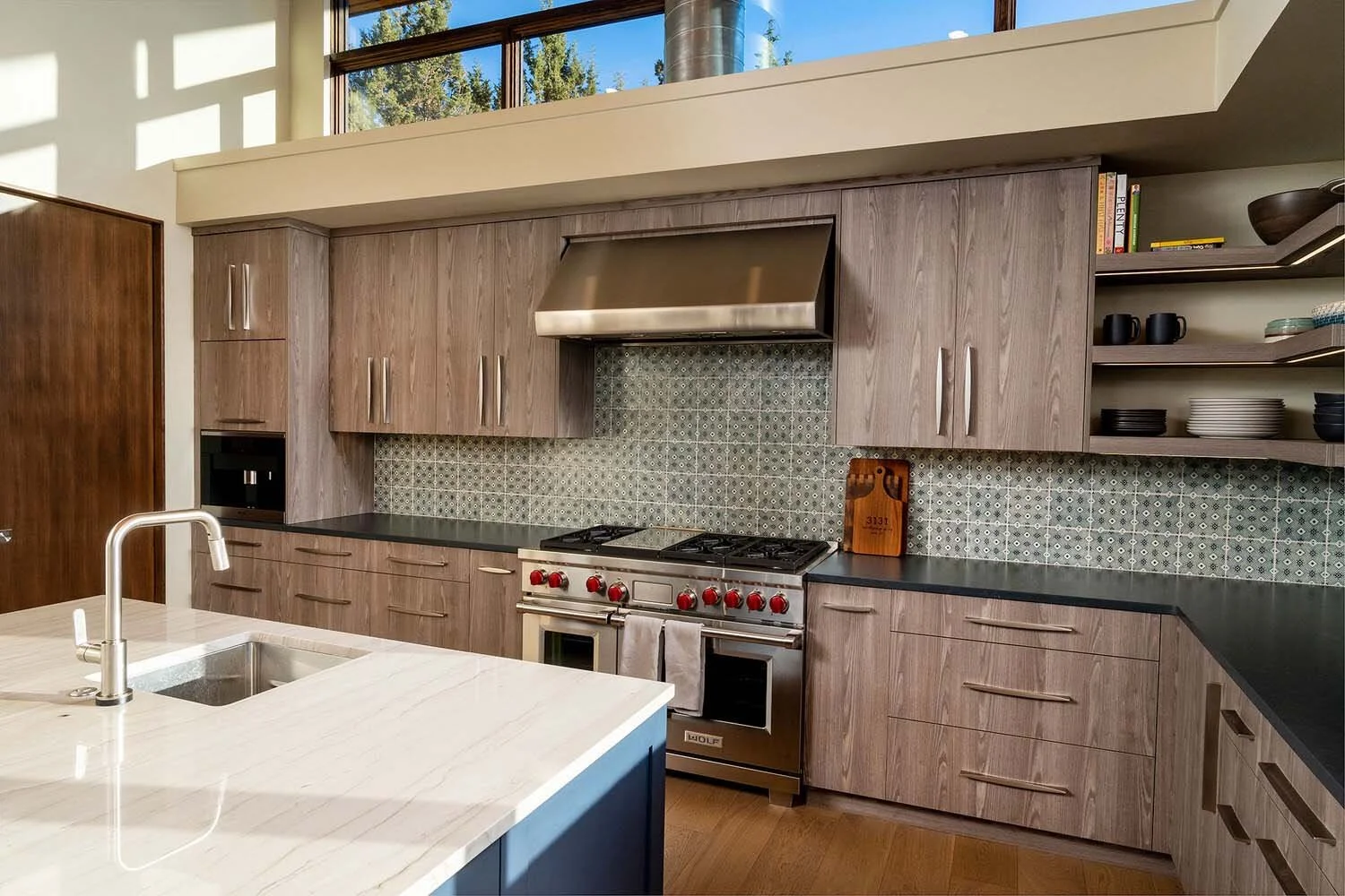 This modern kitchen in Awbrey Butte, a beautiful example of a high-end residential project, features elegant light wood cabinets paired with sleek black countertops and a distinctive patterned backsplash. A professional stainless steel stove anchors