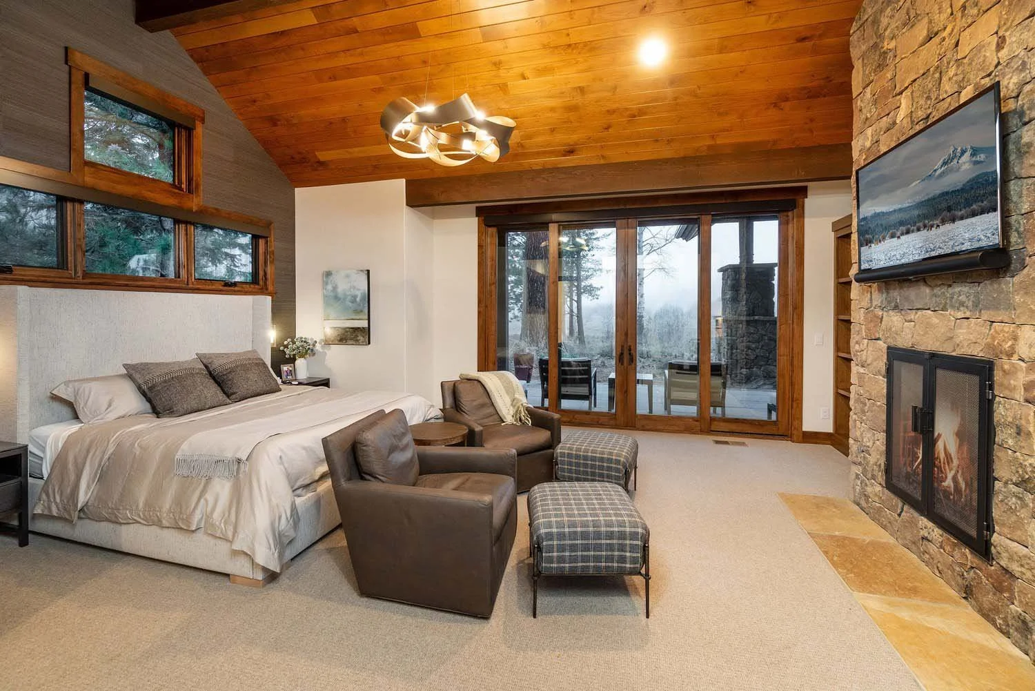 Discover serenity in this inviting bedroom, a hallmark of our luxury custom home designs by Pacwest Builders. A grand bed and two plush brown armchairs with ottomans create a cozy retreat, complemented by a striking stone fireplace crowned with a tel