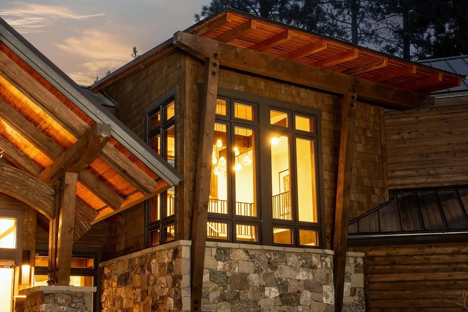 A striking modern house exterior by Pacwest Builders, showcasing a harmonious blend of stone and wood siding under a distinctive roof structure. Large glass windows reflect the sunset, revealing the warm interior lights of this luxury custom home. Th