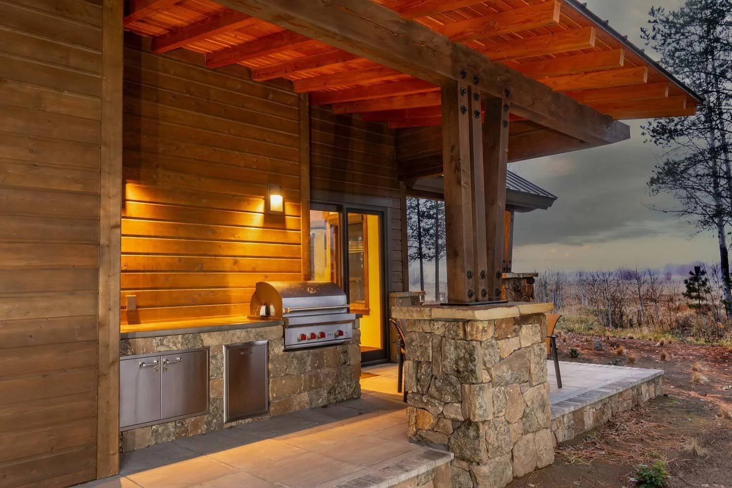 An outdoor kitchen at dusk in Black Butte Ranch, a perfect blend of wood and stone. This luxury custom home features a built-in stainless steel grill and a spacious porch, ideal for enjoying the stunning Central Oregon scenery. Designed and built by
