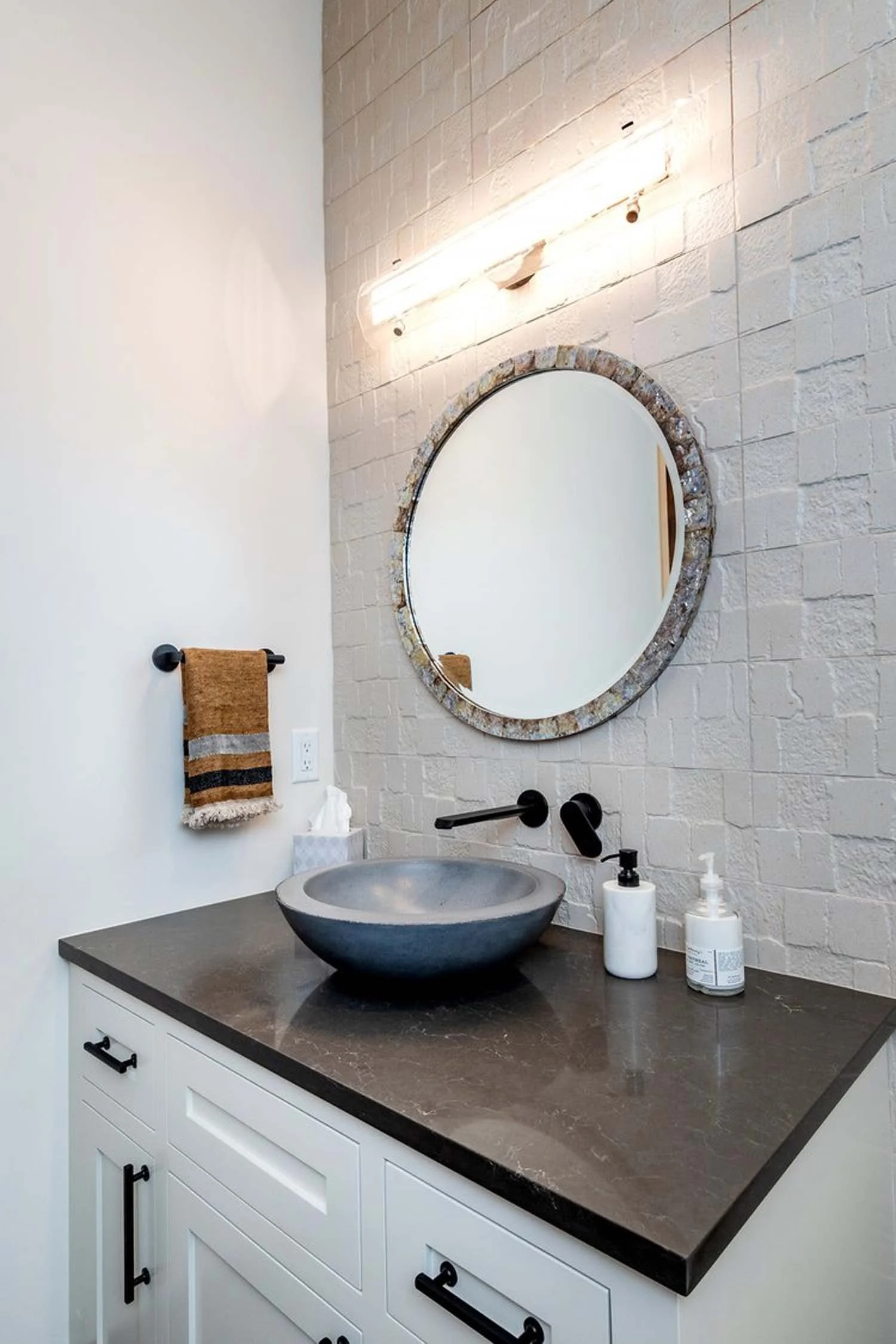 An embodiment of refined elegance, this modern bathroom vanity, expertly crafted by Pacwest Builders, features a dark stone countertop paired with a striking round stone vessel sink. A sleek, wall-mounted black faucet and a circular stone-framed mirr