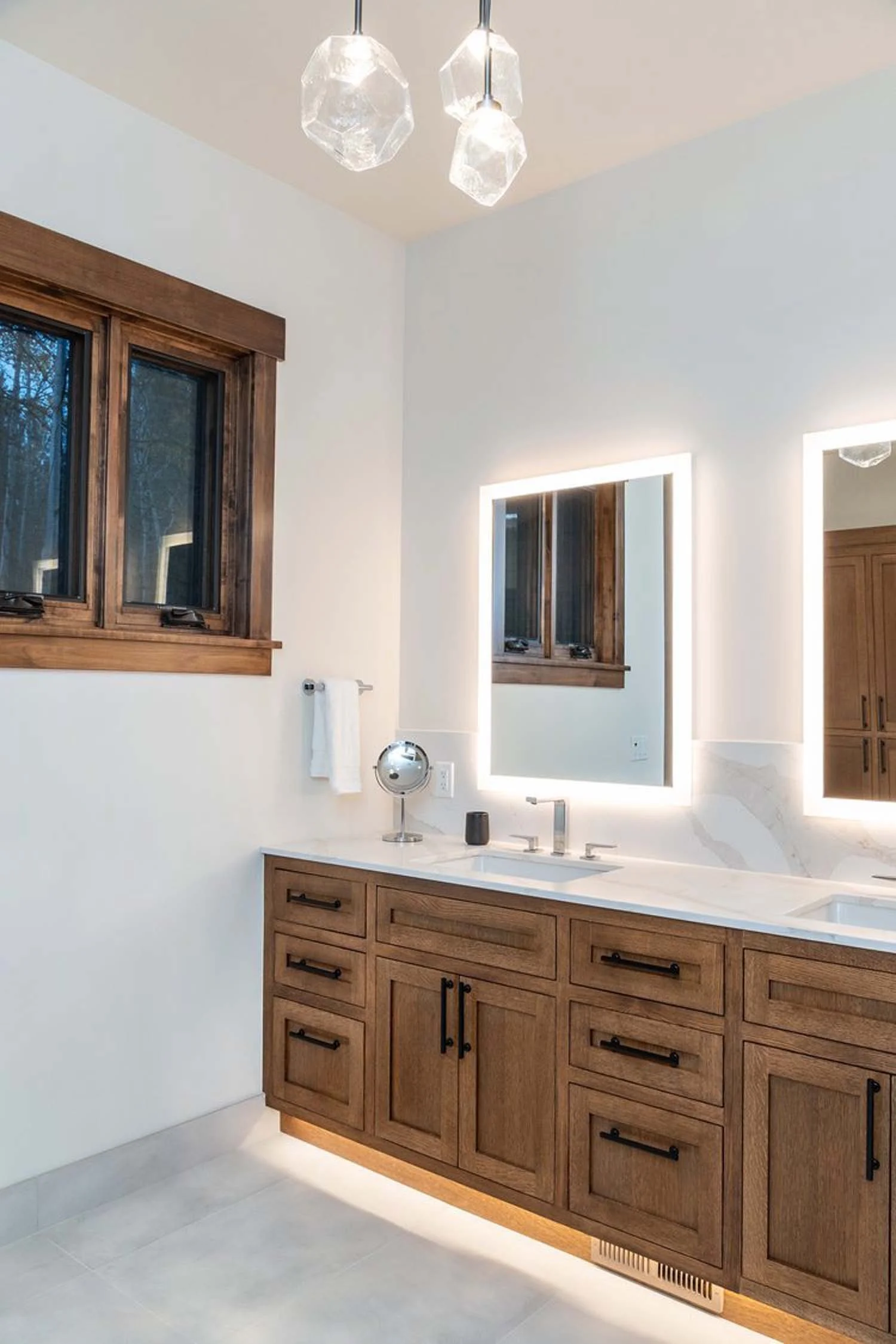 This modern bathroom, designed by Pacwest Builders, showcases exquisite craftsmanship for luxury custom homes. A sleek wooden vanity is perfectly complemented by illuminated mirrors and a window with sophisticated wooden trim, creating a bright and e