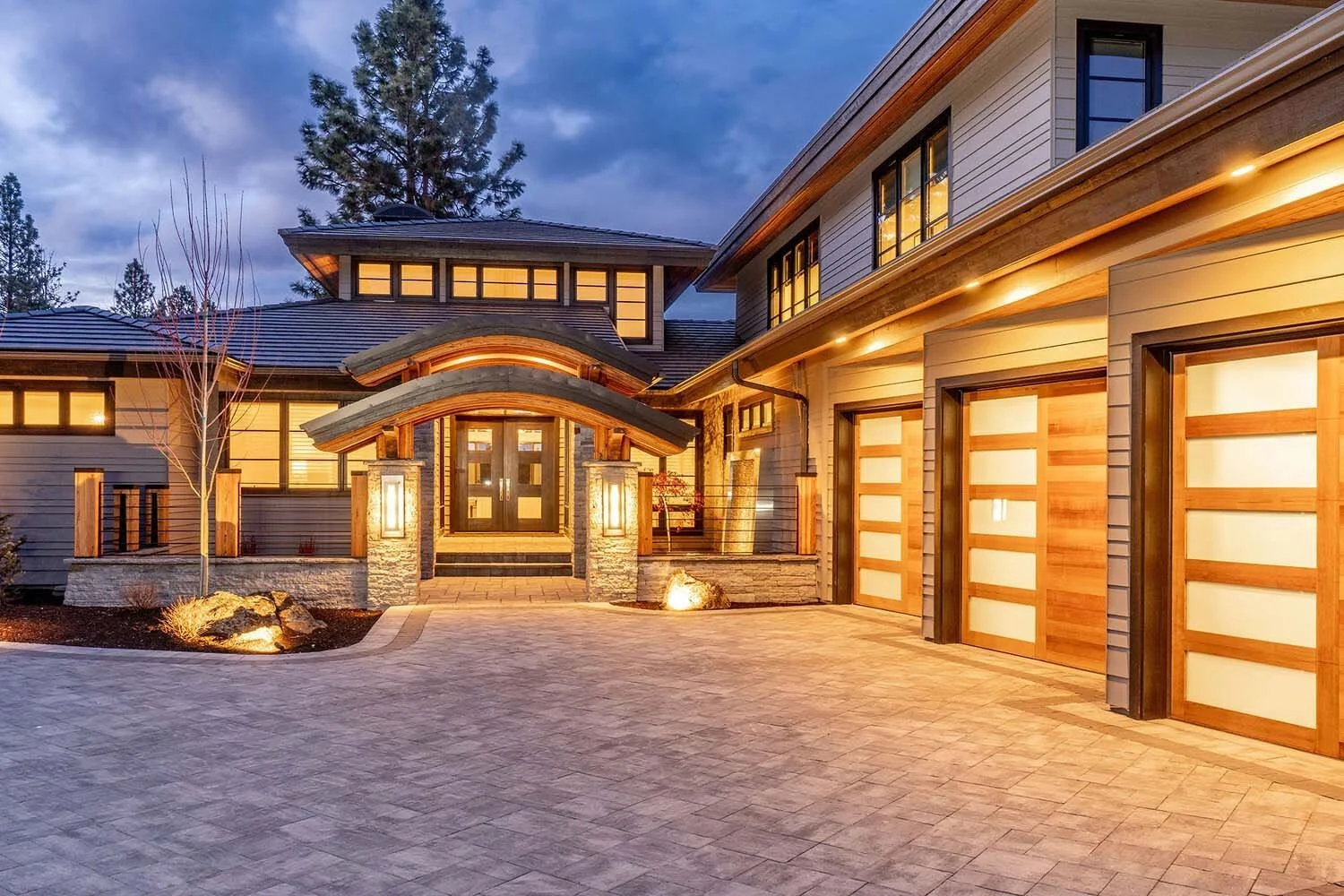 The striking exterior of this luxury custom home in Broken Top designed by Steve Van Sant showcases the refined style of Pacwest Builders. A harmonious mix of stone and wood accents, illuminated by outdoor lighting in the evening, creates an inviting