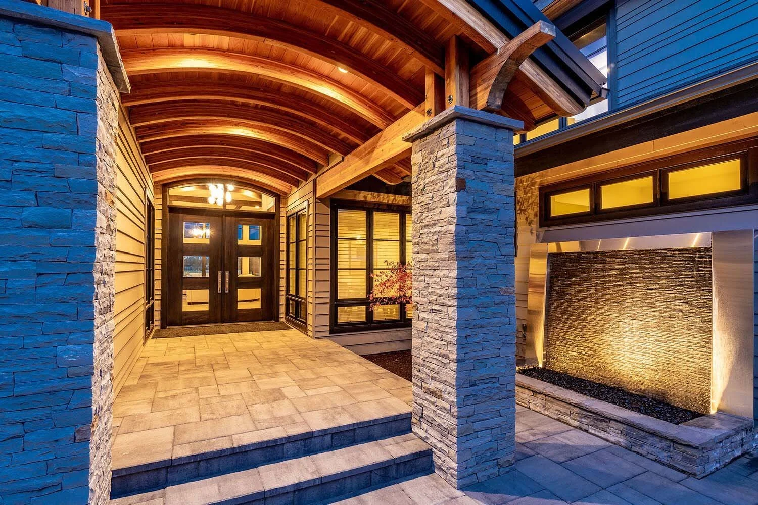 Step into elegance: this modern entrance, a Pacwest Builders luxury custom home masterpiece, features a stunning wooden curved ceiling and stately stone columns. Grand glass double doors welcome you, while a beautifully lit water feature adds serene