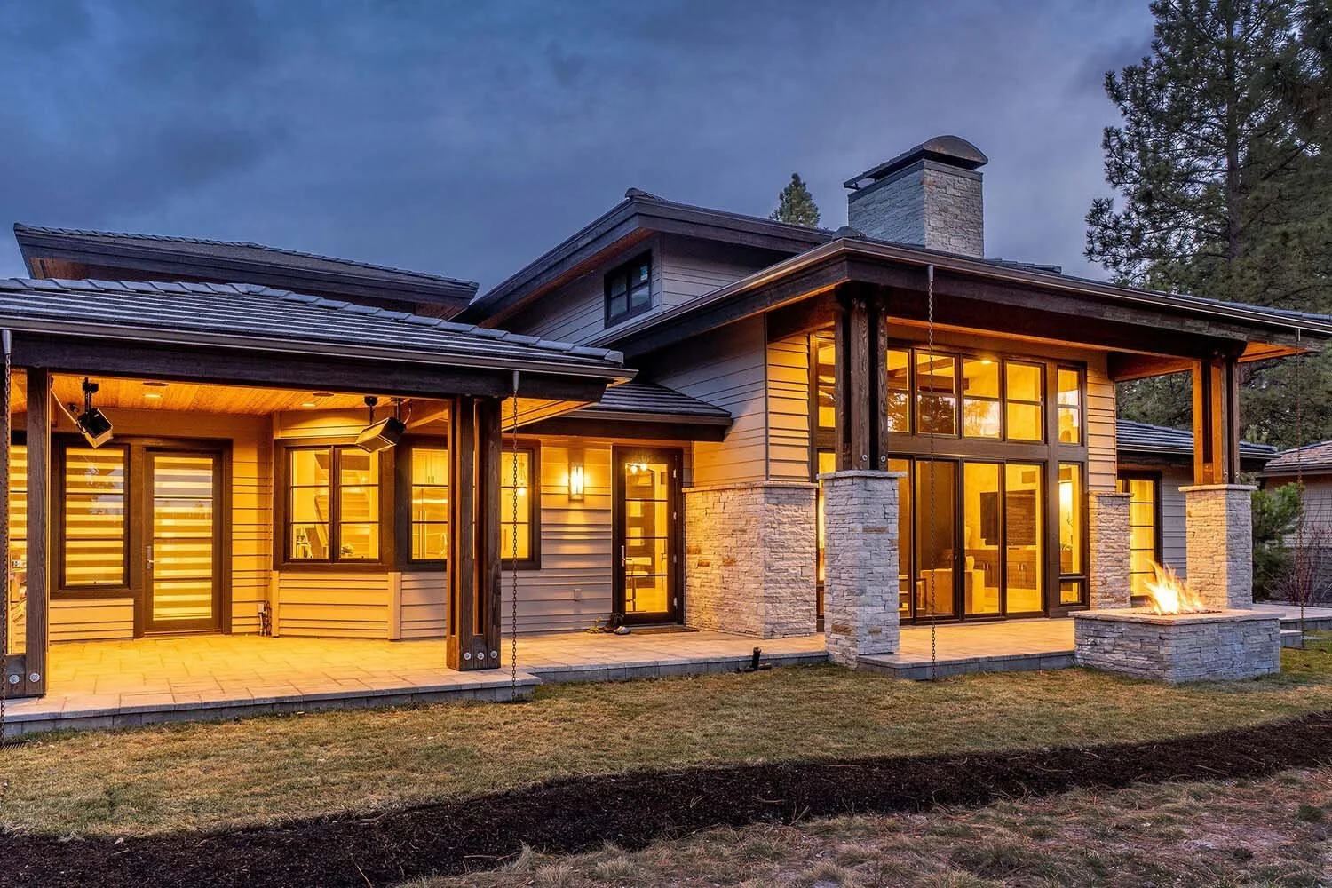 As dusk settles over Broken Top, this luxury custom home by Pacwest Builders comes alive. A expansive patio, framed by illuminated windows, invites relaxation around a captivating fire feature. Surrounded by the tranquility of mature trees, this outd
