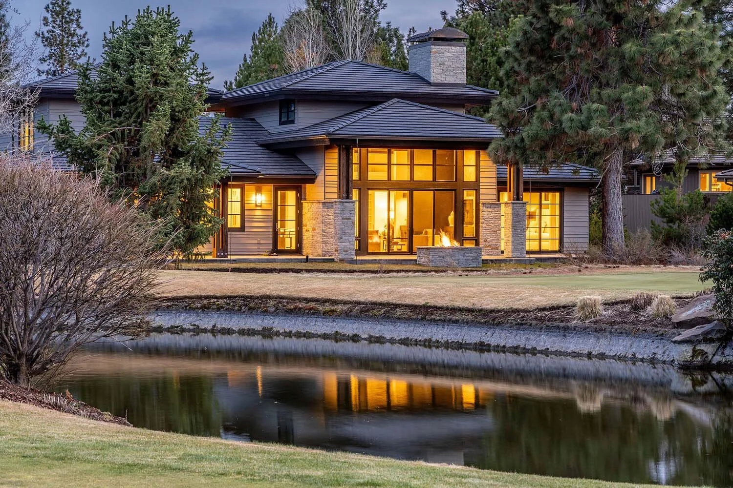 Witness the captivating beauty of this modern luxury custom home by Pacwest Builders at dusk. Expansive glass windows and a prominent stone fireplace illuminate the serene landscape, reflecting beautifully on the nearby pond. Surrounded by lush trees