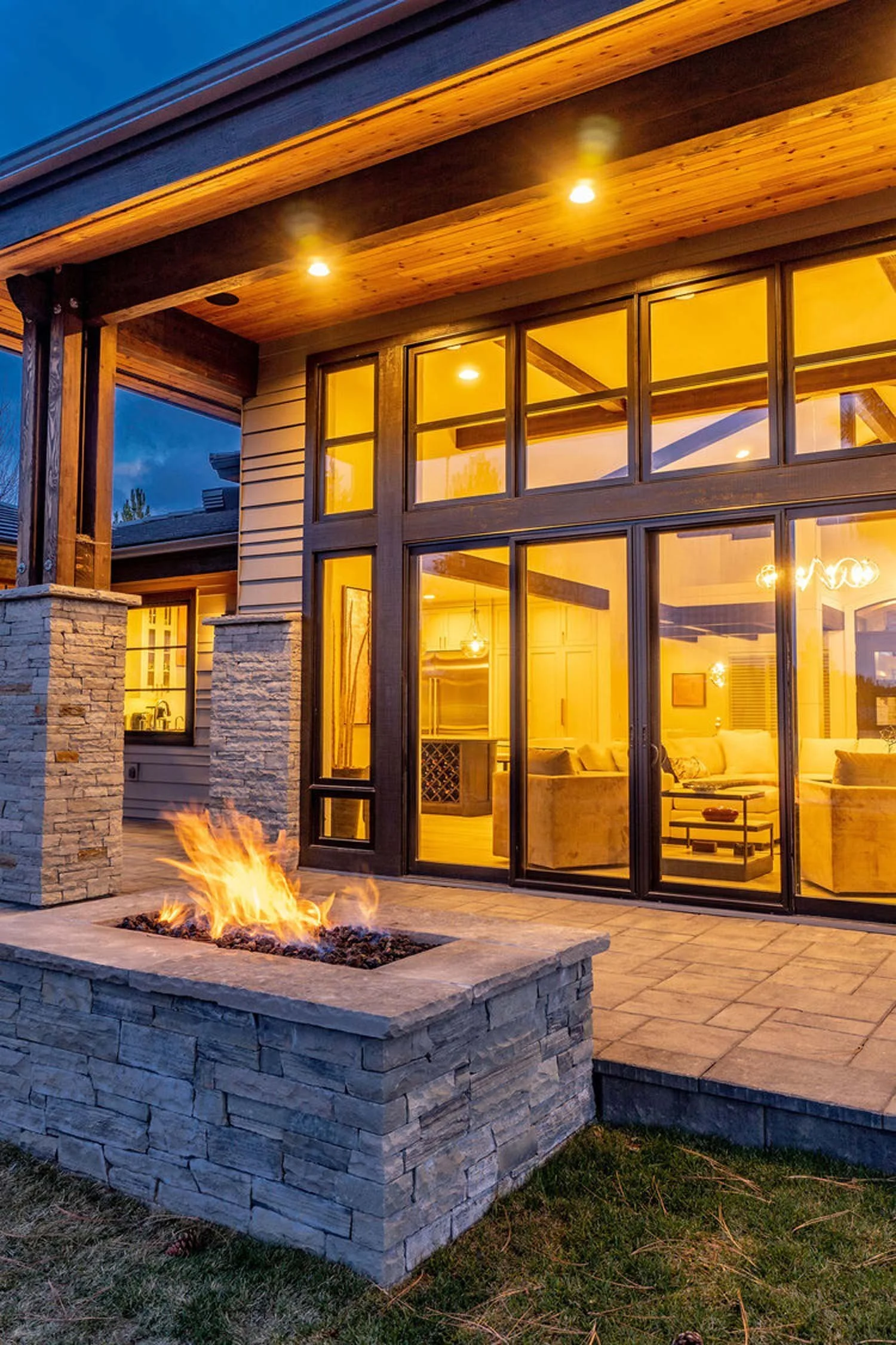 This striking exterior view captures a modern luxury custom home by Pacwest Builders, where expansive glass windows invite warm yellow light to spill out, creating an inviting glow at dusk. A beautifully crafted stone firepit anchors the outdoor livi