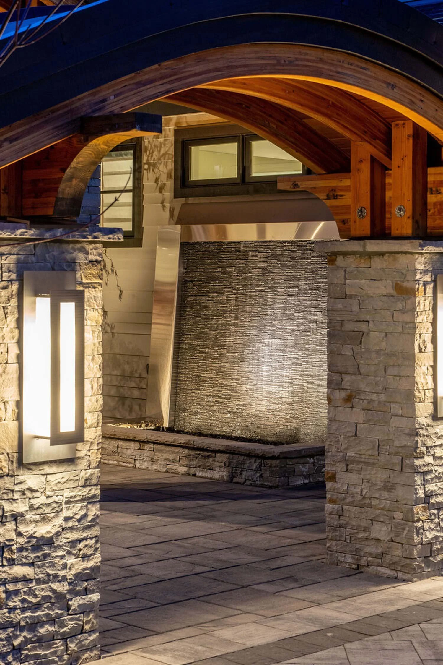 Behold this stunning nighttime view of a modern outdoor architectural feature, a testament to Pacwest Builders' innovative design in luxury custom homes. Robust stone pillars frame a captivating, textured waterfall wall, beautifully complemented by e