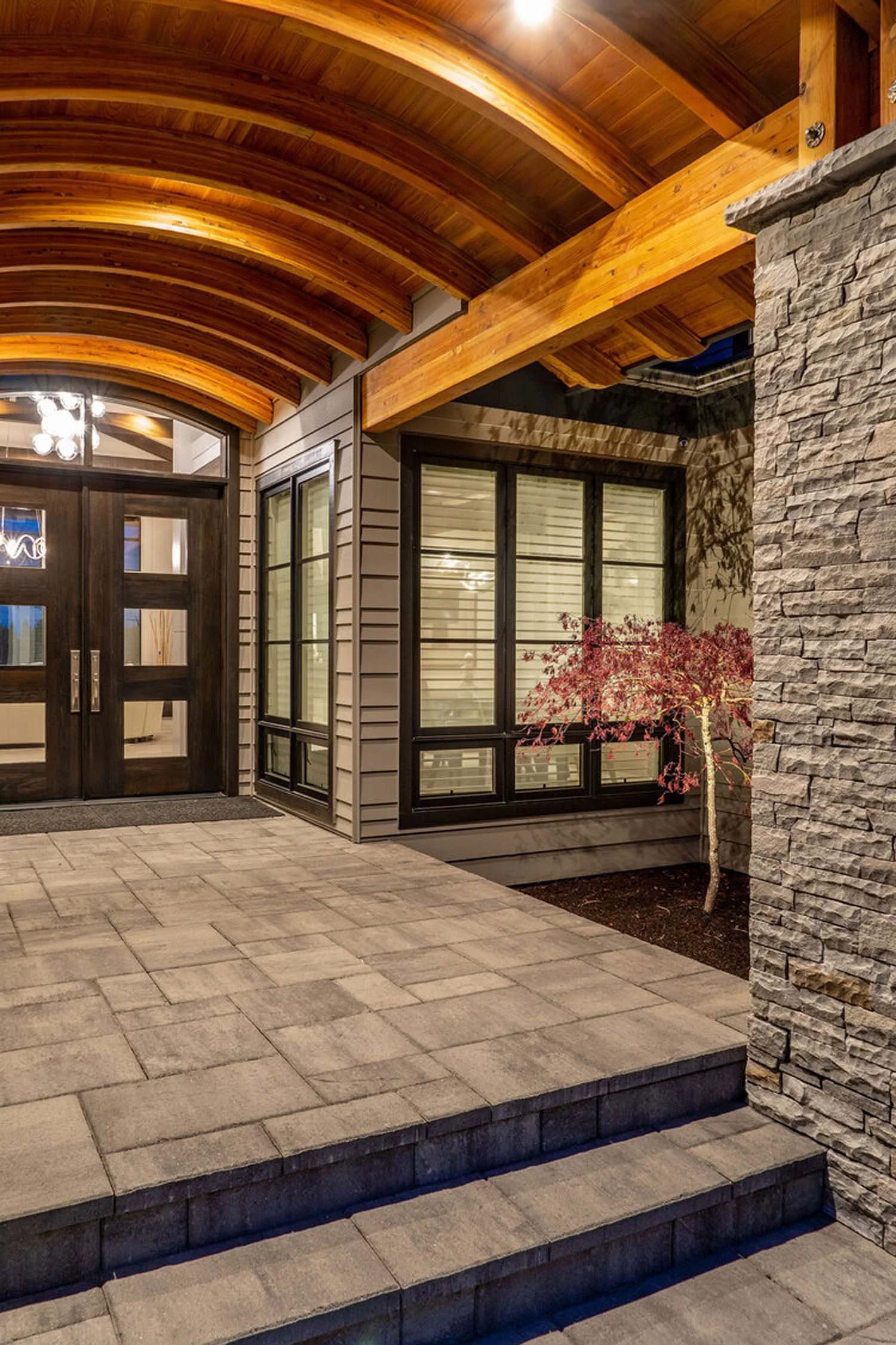This enchanting night view reveals the exterior porch of a modern luxury custom home by Pacwest Builders, where elegant stone and warm wood accents converge. A small tree with vibrant red leaves adds a touch of natural artistry, while large windows g