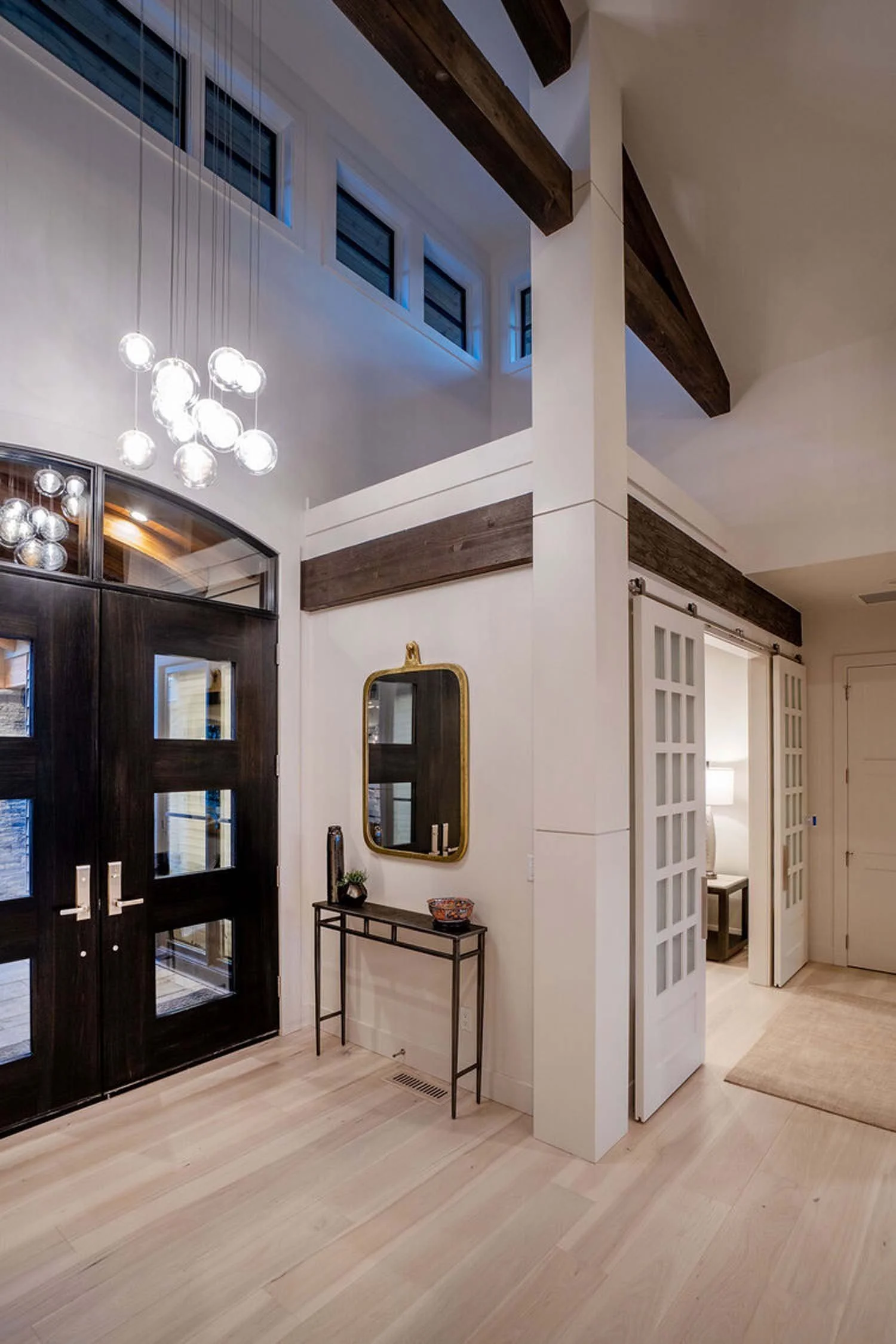This elegant entryway, showcasing exquisite interior design by Kristi Yozamp, is a highlight in a luxury custom home by Pacwest Builders. Black double doors open to reveal white walls, a striking gold-framed mirror above a small black table with deco