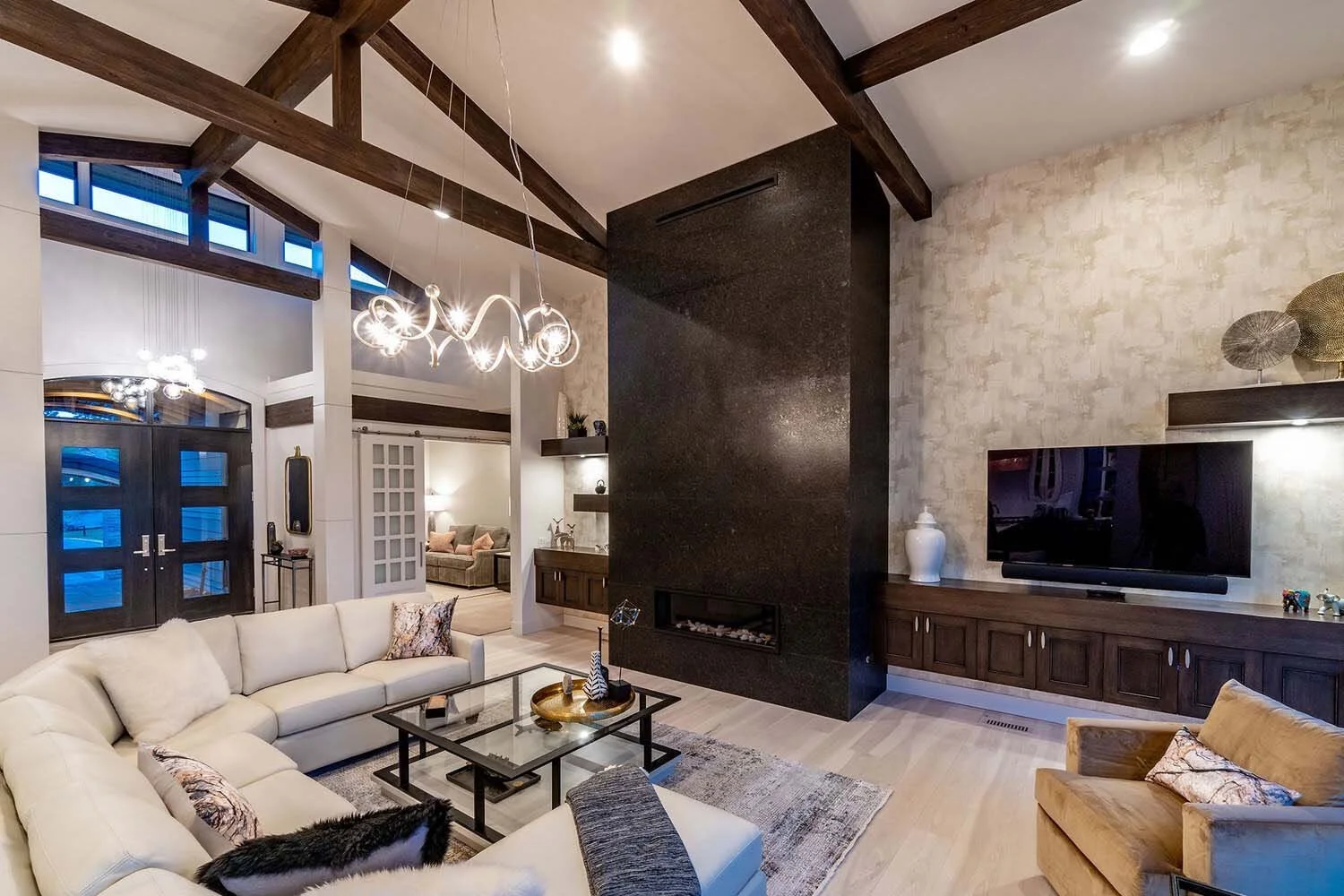 This stunning living room, a masterpiece in a luxury custom home by Pacwest Builders, captivates with its high vaulted ceiling and majestic exposed wooden beams. A grand black fireplace commands attention, complemented by a luxurious cream sectional
