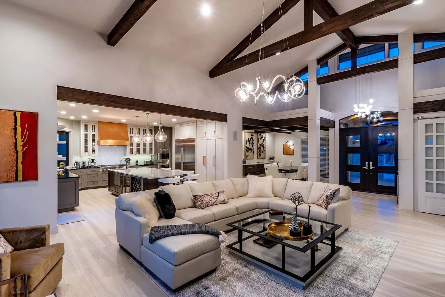 This expansive, open-concept living room, flowing seamlessly into the kitchen, exemplifies the innovative design of luxury custom homes by Pacwest Builders. Majestic exposed beams define the space, complemented by unique decorative light fixtures tha