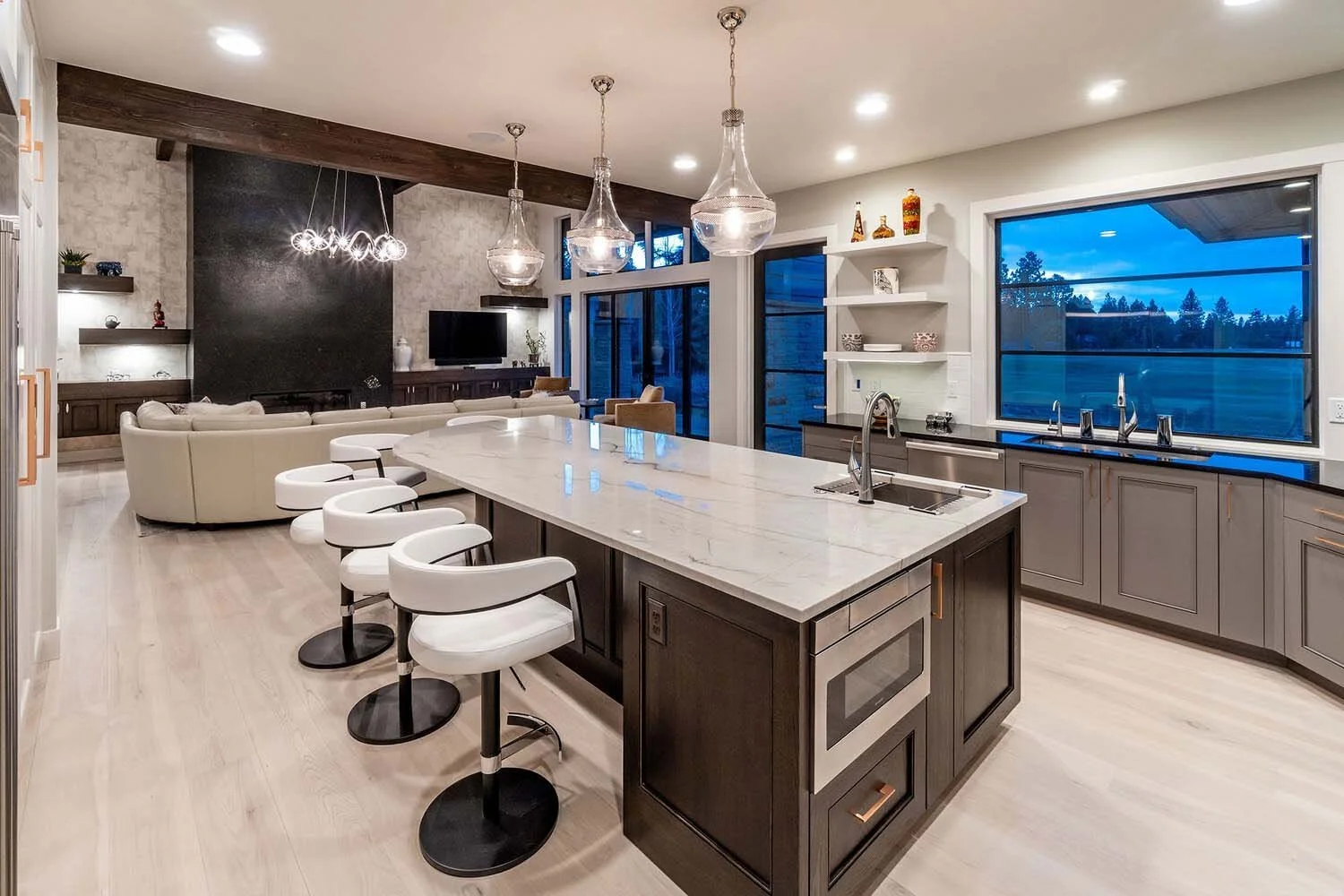 This vibrant open-concept kitchen and living room, a showcase of modern decor in a luxury custom home by Pacwest Builders, features a sleek island with a stunning marble countertop. White and black bar stools add a contemporary touch, while large win