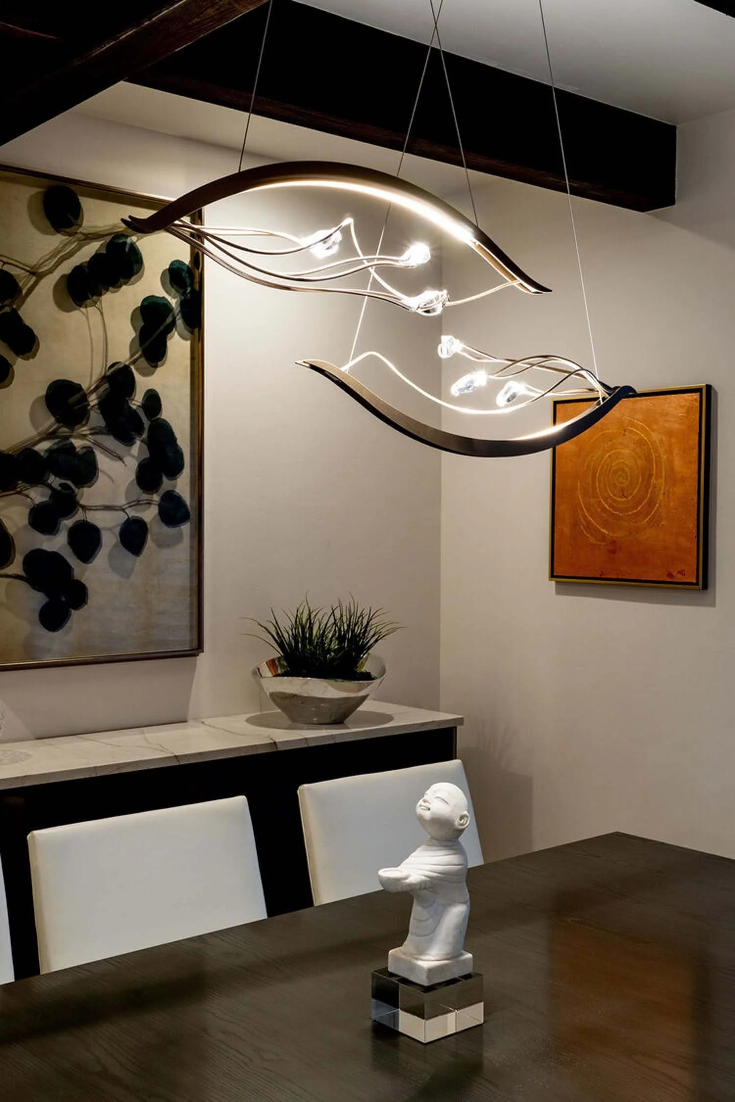 This sophisticated modern dining room, a highlight in a luxury custom home by Pacwest Builders, features a sleek dark wood table paired with elegant white chairs. Abstract artwork adorns the walls, while a decorative plant on a stylish sideboard adds