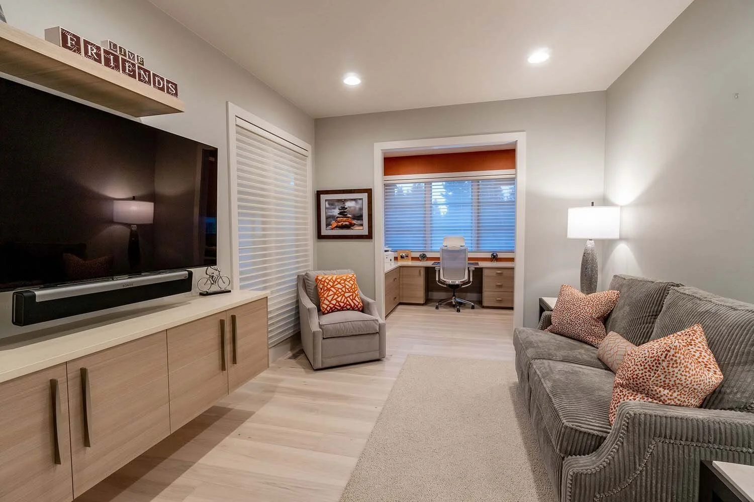 This inviting living room, a hallmark of Pacwest Builders' luxury custom homes, offers both comfort and style. Featuring a discreetly placed TV on a beige cabinet, it includes a cozy gray armchair with an orange accent pillow and a complementary gray