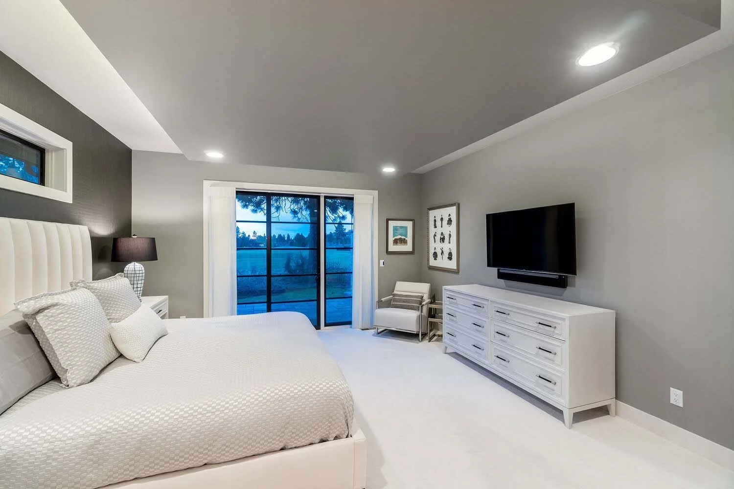 This serene modern bedroom, a testament to Pacwest Builders' luxury custom homes, offers a tranquil retreat. Featuring a crisp white bed, a convenient bedside lamp, and a wall-mounted TV for discreet entertainment, it combines comfort with contempora