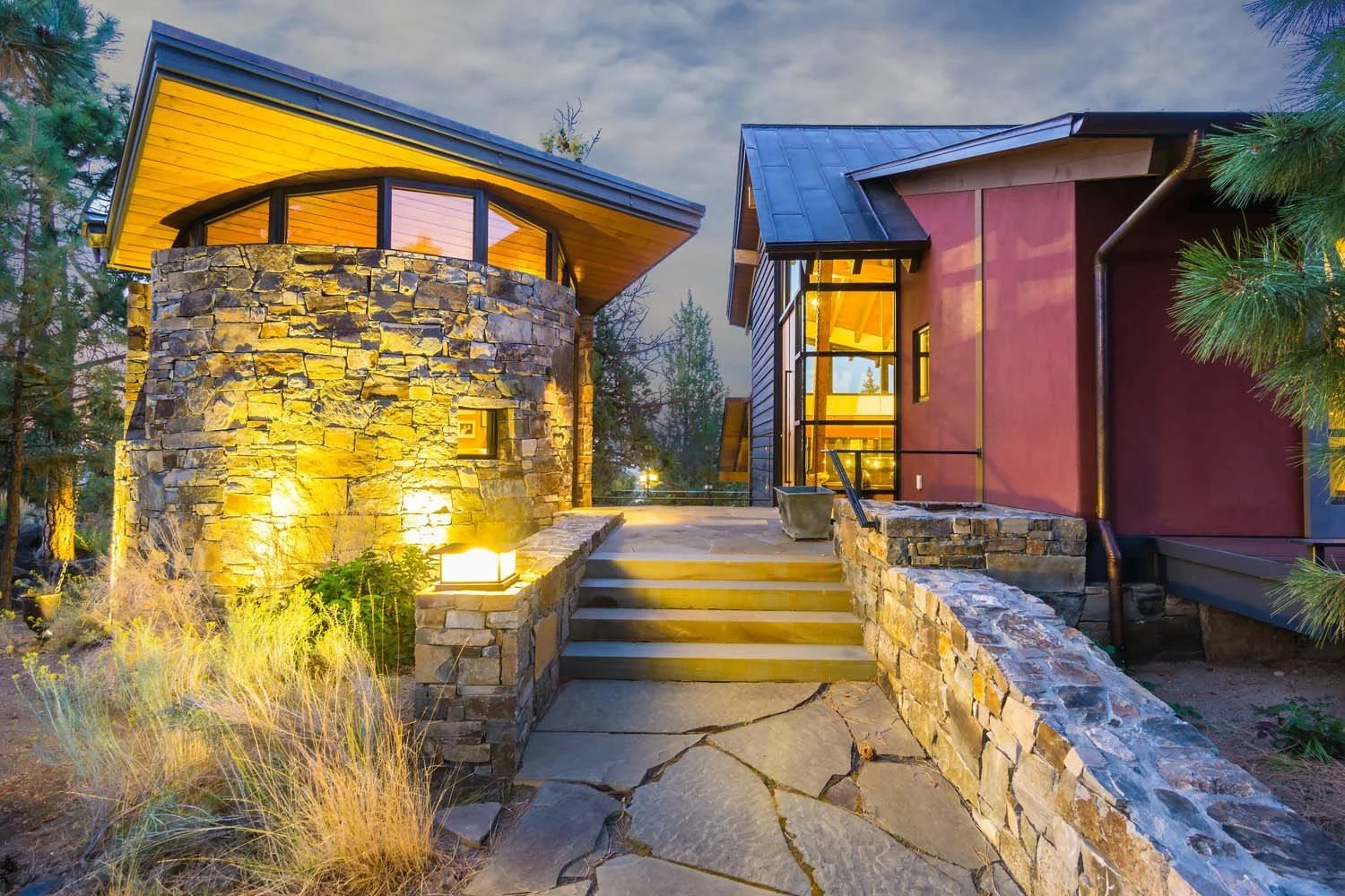 This captivating modern home in North Rim - Puccoon Court, a Hayes Inc. design and Pacwest Builders' construction, showcases a harmonious stone and wood exterior, beautifully illuminated by outdoor lighting. Surrounded by lush trees and plants, a wel