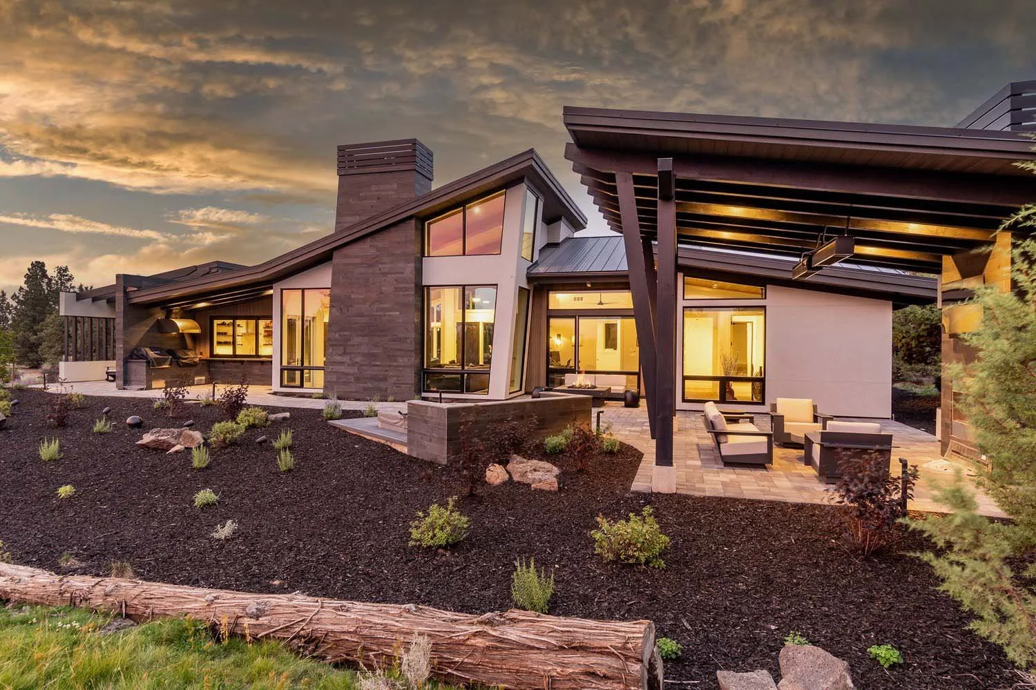 This peaceful view of the Pronghorn - Canyon View home, designed by Blair Davis, showcases its perfect harmony with the landscape. The home's expansive large windows reflect the warm glow of sunset, while the inviting outdoor patio furniture offers a