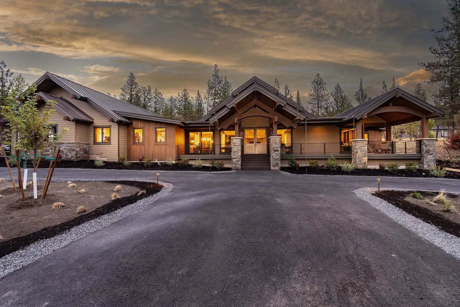 This spectacular front view of the Skyline Ranch - Imbler home, built by Pacwest Builders, showcases its impressive scale and modern design. The home is beautifully set against a backdrop of trees at sunset, with a sleek dark asphalt driveway leading