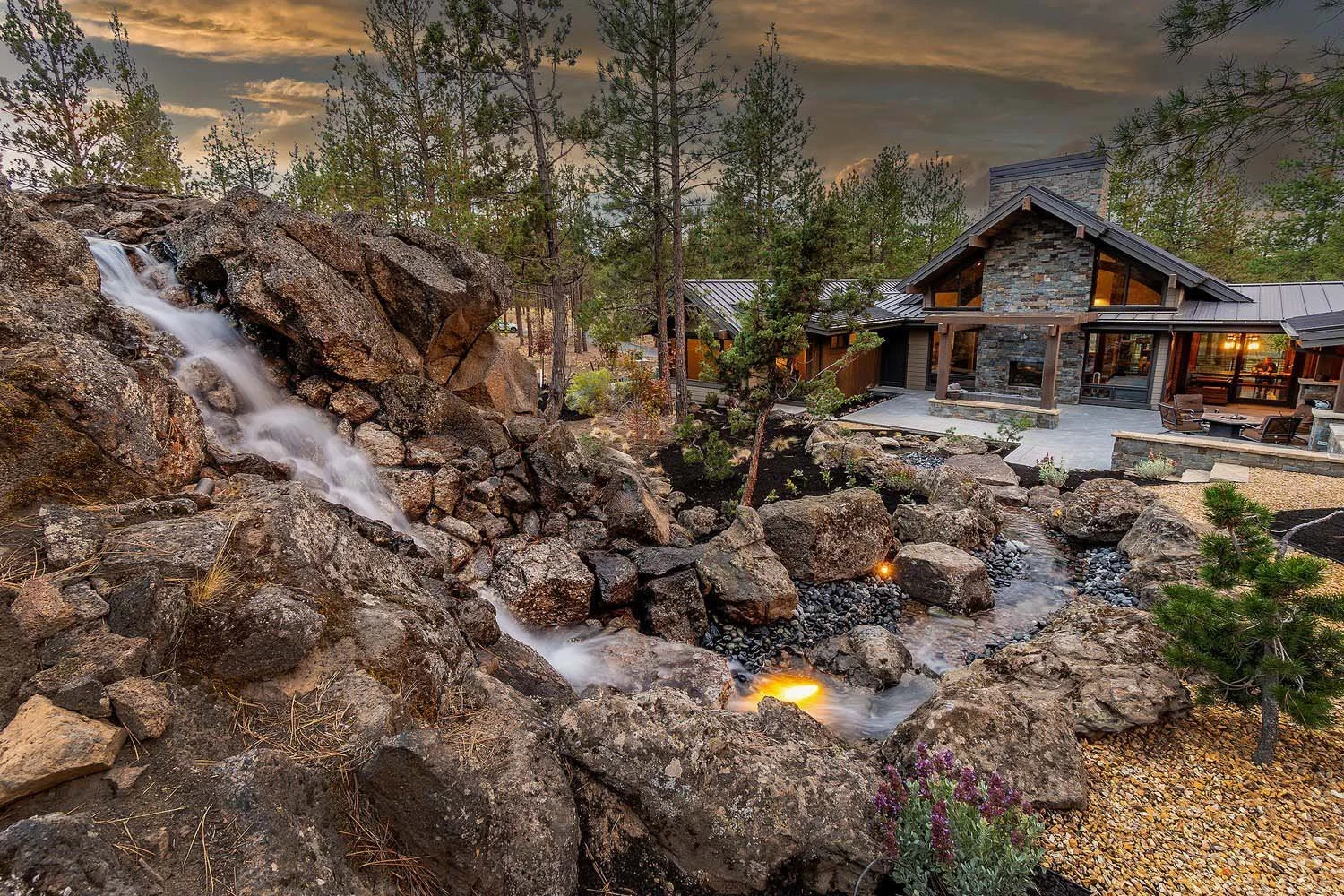 This spectacular modern house at Skyline Ranch - Imbler, built by Pacwest Builders, is beautifully framed by trees against a stunning sunset sky. The home features expansive large windows and a prominent stone chimney. In the foreground, a tranquil r