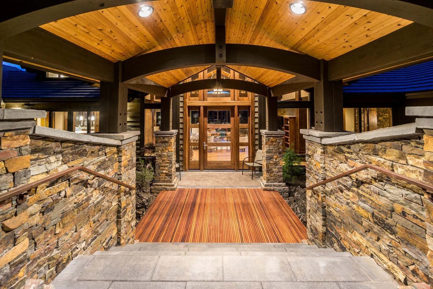 The stunning entrance of this award-winning luxury custom home at North Rim - Wild Rye makes a dramatic statement. A beautifully designed walkway with stone walls and wooden elements leads to a sleek glass door, all under a richly colored wooden deck
