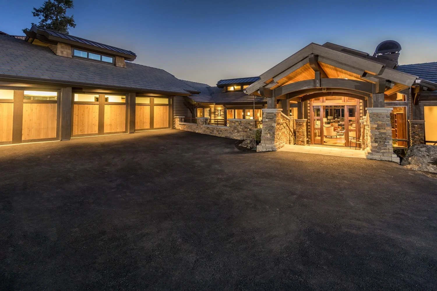 This stunning front view of the award-winning North Rim - Wild Rye home, built by Pacwest Builders, showcases its impressive scale and design. The home features a harmonious wooden and stone exterior, complemented by expansive large windows that glow
