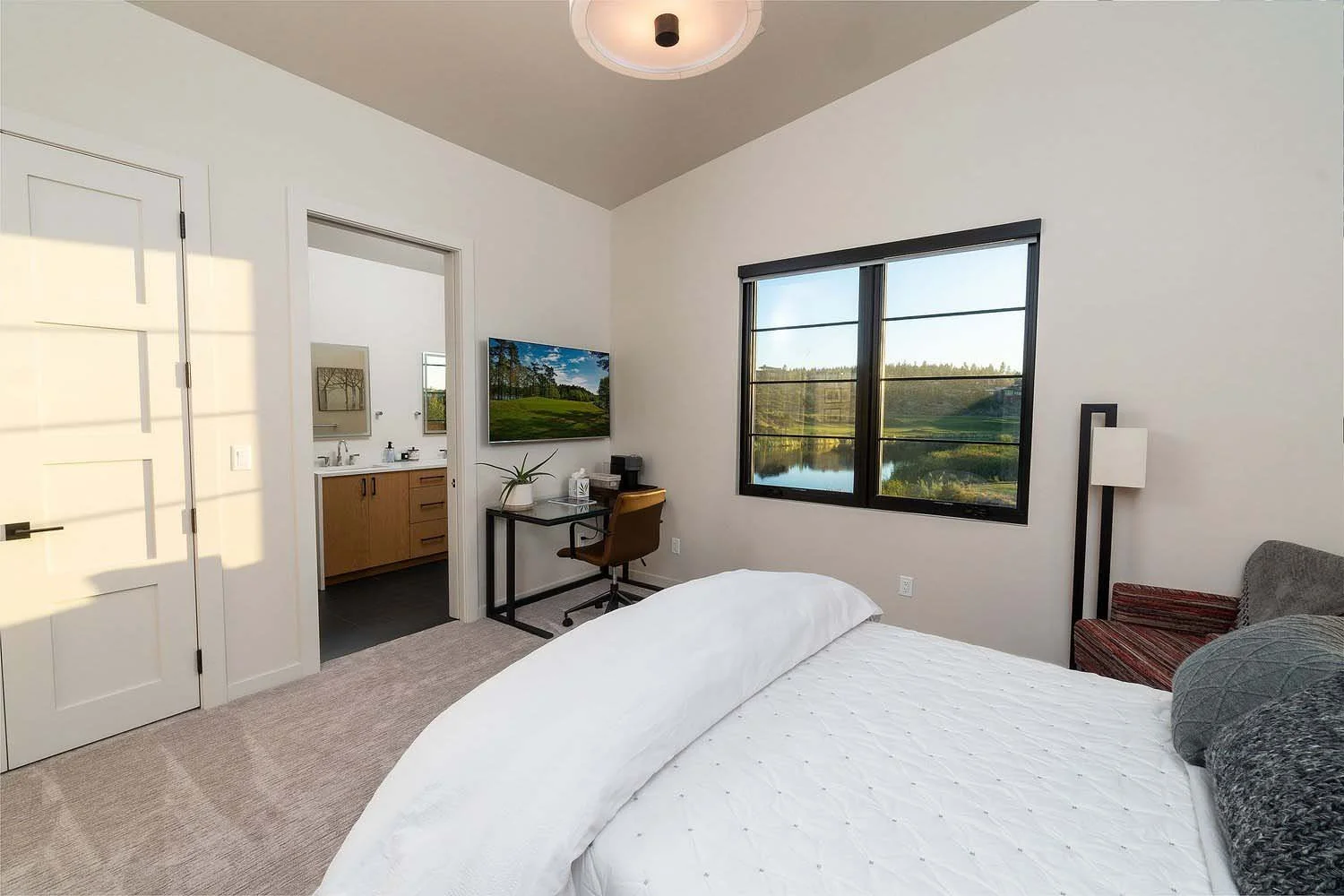 This peaceful bedroom at the Tetherow - Hardin Martin Court home by Pacwest Builders is a true retreat designed to connect seamlessly with the outdoors. The space features a cozy bed with white bedding and a small workspace with a TV. The highlight i
