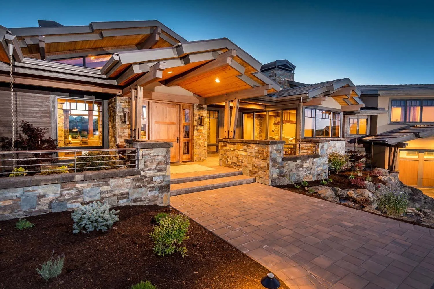 Pacwest Builders masterfully designed this stunning mountain modern luxury custom home in Central Oregon, at Tetherow - Hawk Court. This image highlights the home’s striking architecture, including a beautiful stone and wood exterior. The brick pathw