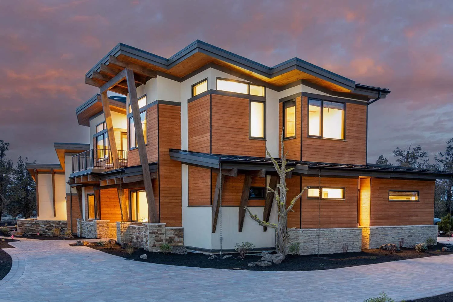 Luxury custom multi-story home in Pronghorn - Switchback, built by Pacwest Builders. This modern Central Oregon residence, designed by Blair Davis, features a striking wood and white exterior. The home's large, illuminated windows and paved pathway c