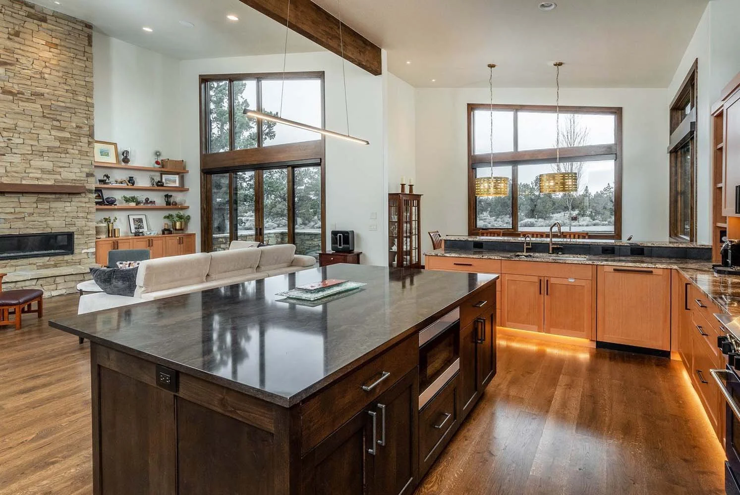 This open-concept kitchen and living room showcases the impeccable architecture of a luxury custom home in Pronghorn - Switchback. The space, built by Pacwest Builders, features large windows that highlight the beautiful wood flooring and a stunning