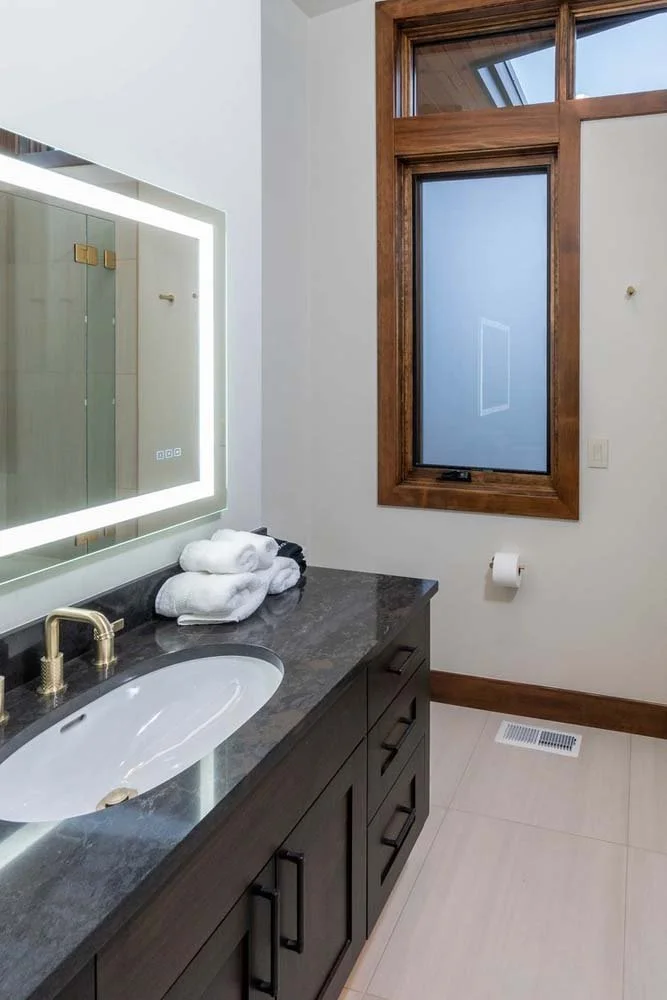 This modern bathroom in Pronghorn - Switchback by Pacwest Builders showcases stunning interior design. The space features a sleek dark countertop with a white sink and a large illuminated mirror, complemented by a window with wooden trim. This luxury