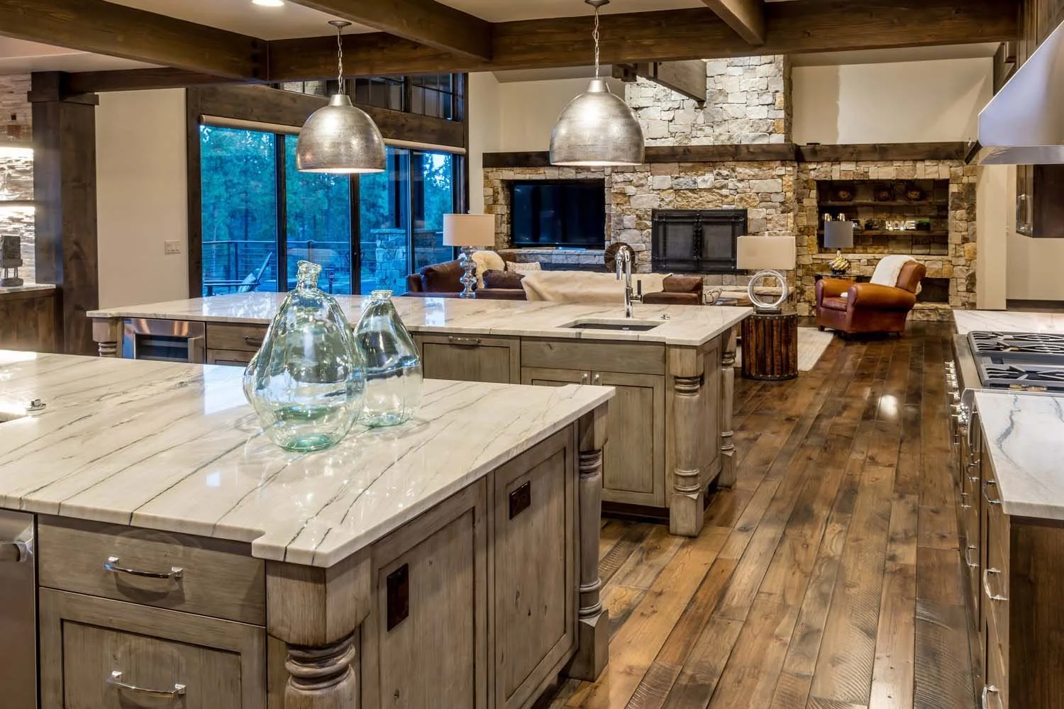 This open-concept kitchen and living room in Tetherow - Bainbridge Court is a prime example of a mountain modern residence with a rustic feel. The space features a large kitchen island with glass vases and a stone fireplace, creating a cozy and invit
