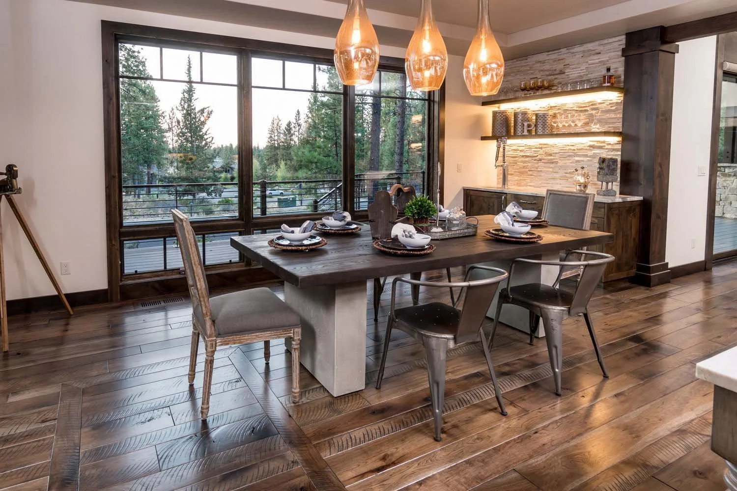 This rustic dining room in Tetherow - Bainbridge Court is a highlight of this mountain modern residence. The space, with its large wooden table and chairs, is set beside floor-to-ceiling windows, showcasing a view of the trees. The architecture by Sp
