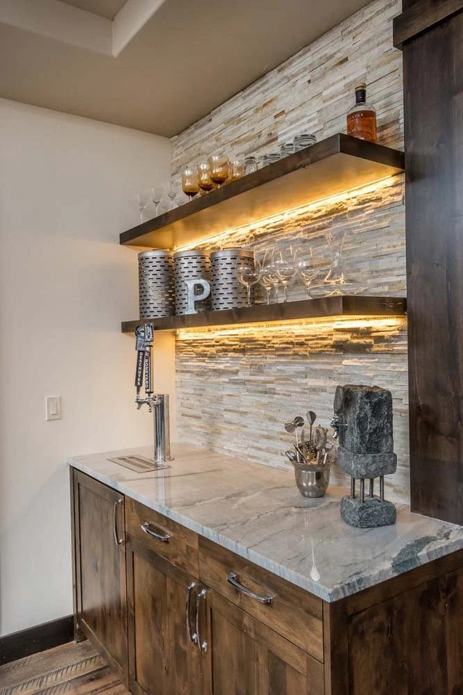 A modern bar area with a marble countertop, wood cabinets, and floating shelves with LED lighting, part of the Tetherow - Bainbridge Court project in Central Oregon.
