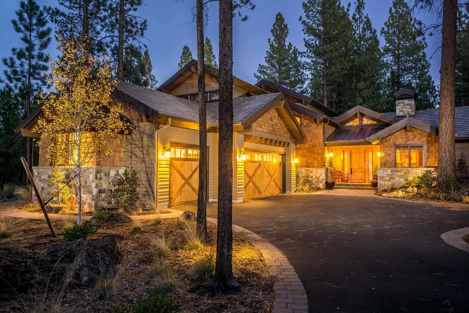 Designed by Spencer Williams, this mountain modern home in Central Oregon won "Best Architectural Design" and "Best of Show" for its category. The image shows the house at night in a wooded area, with warm exterior lights illuminating its stone and w