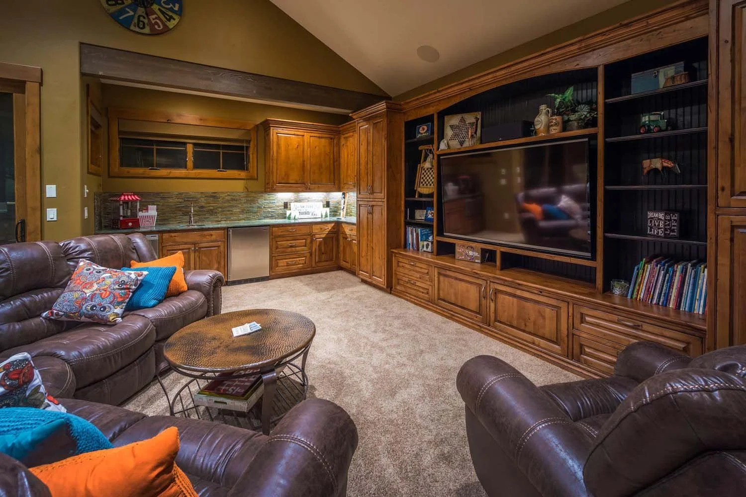 Designed by Spencer Williams, this cozy living room, part of the "Best of Show" and "Best Architectural Design" winning home, features comfortable leather sofas, a wooden entertainment center, and a small kitchenette. The built-in shelves are filled