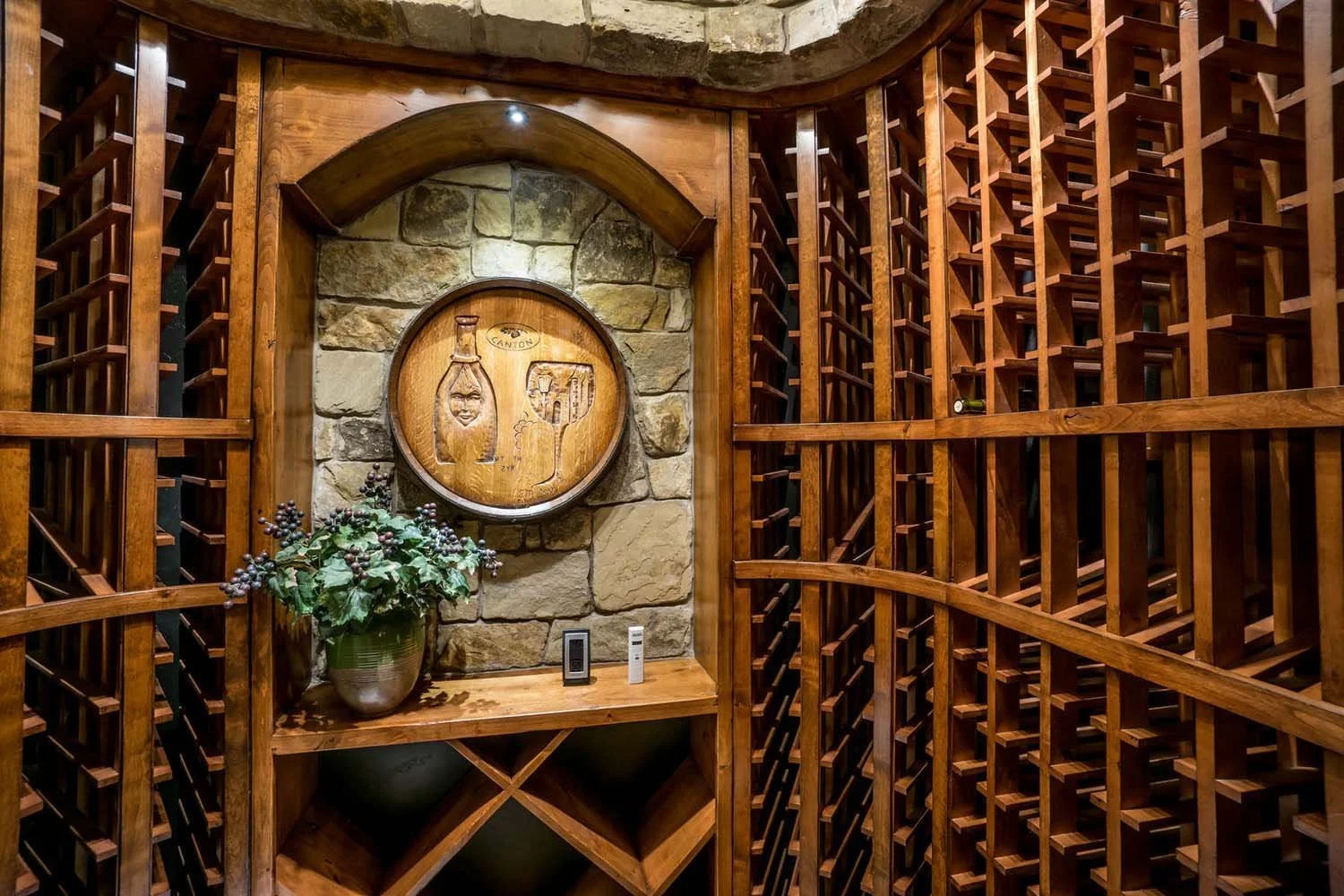 This mountain modern wine cellar, a key feature of the "Best of Show" and "Best Architectural Design" winning home, was designed by Spencer Williams. The space features wooden wine racks, a rustic stone wall, and a unique decorative plaque, perfect f
