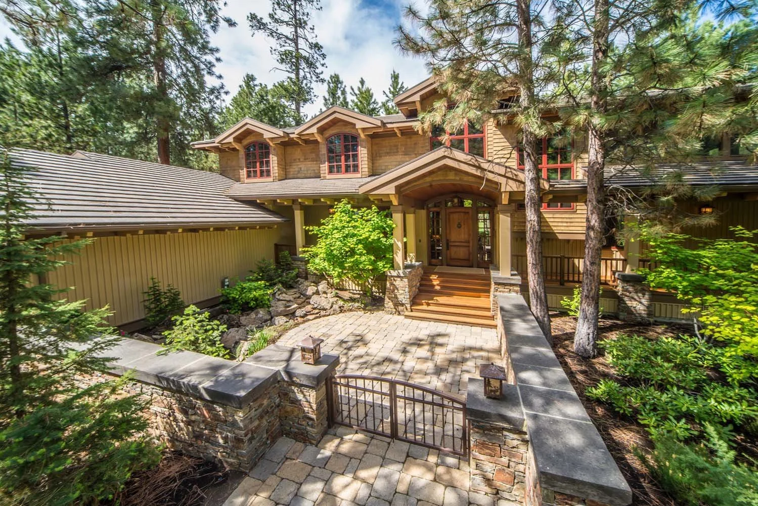 This luxury custom home in Black Butte Ranch - Foin Follette showcases masterful architecture design by Steve Van Sant. The large house features a rustic stone and wood exterior, surrounded by trees and greenery, leading to a beautiful wooden front d